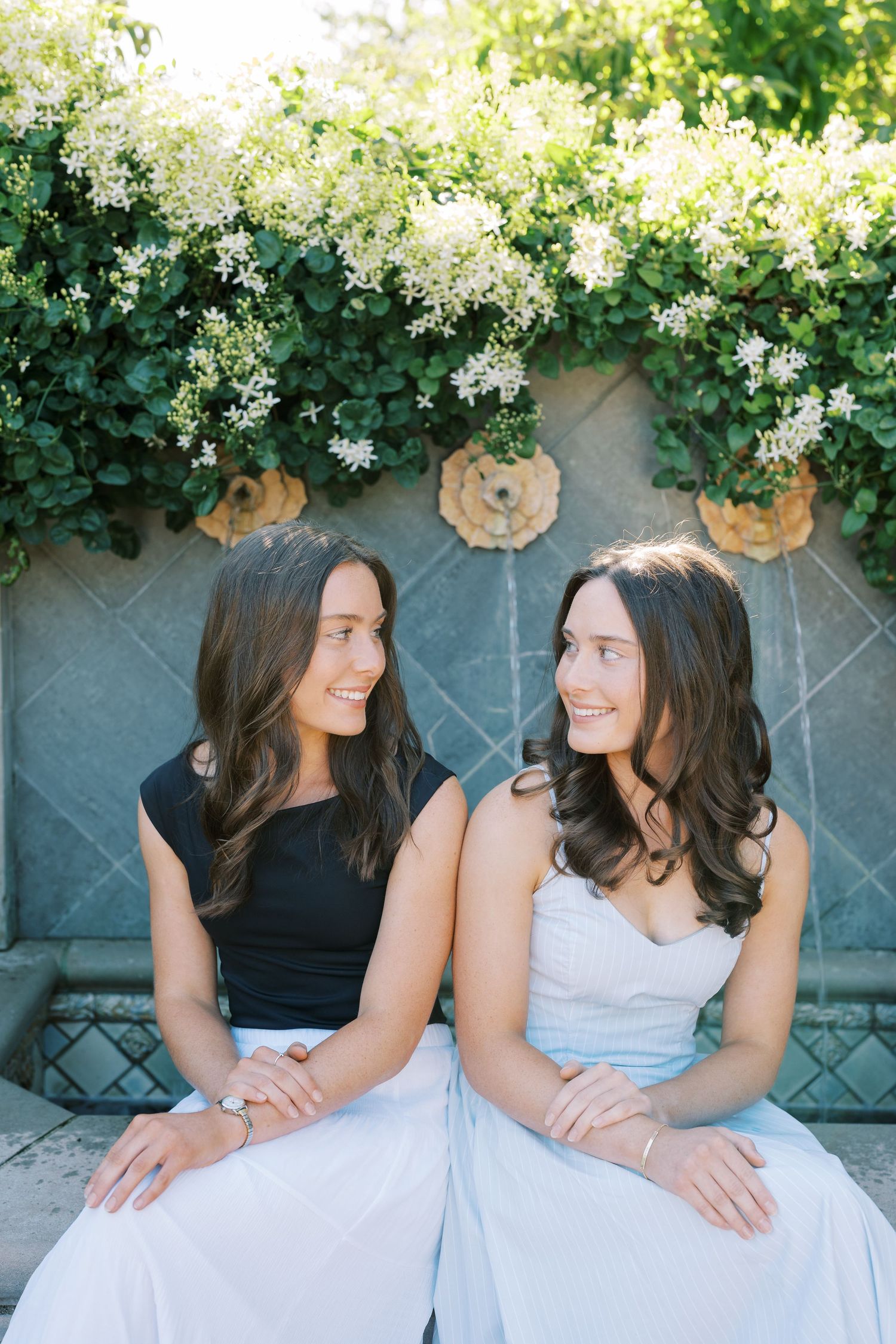 Two people in contrasting outfits sitting close together on a stone bench surrounded by flowering bushes.