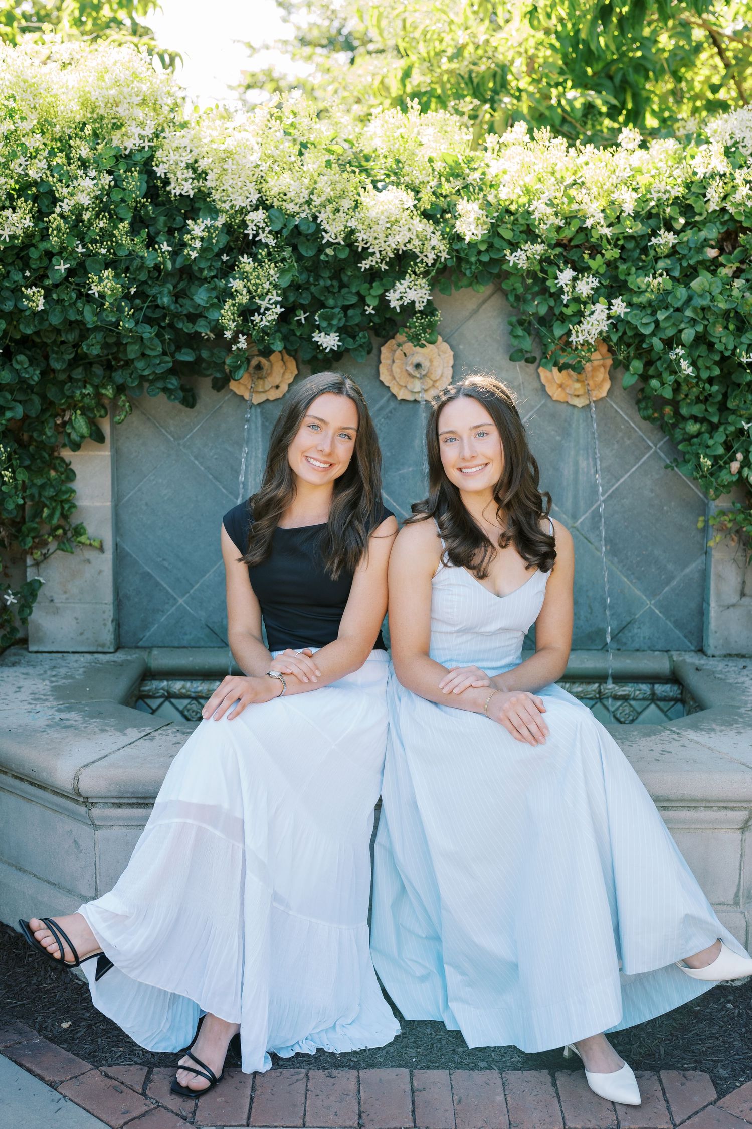 Two people in matching white dresses sit together on stone steps surrounded by lush green foliage.