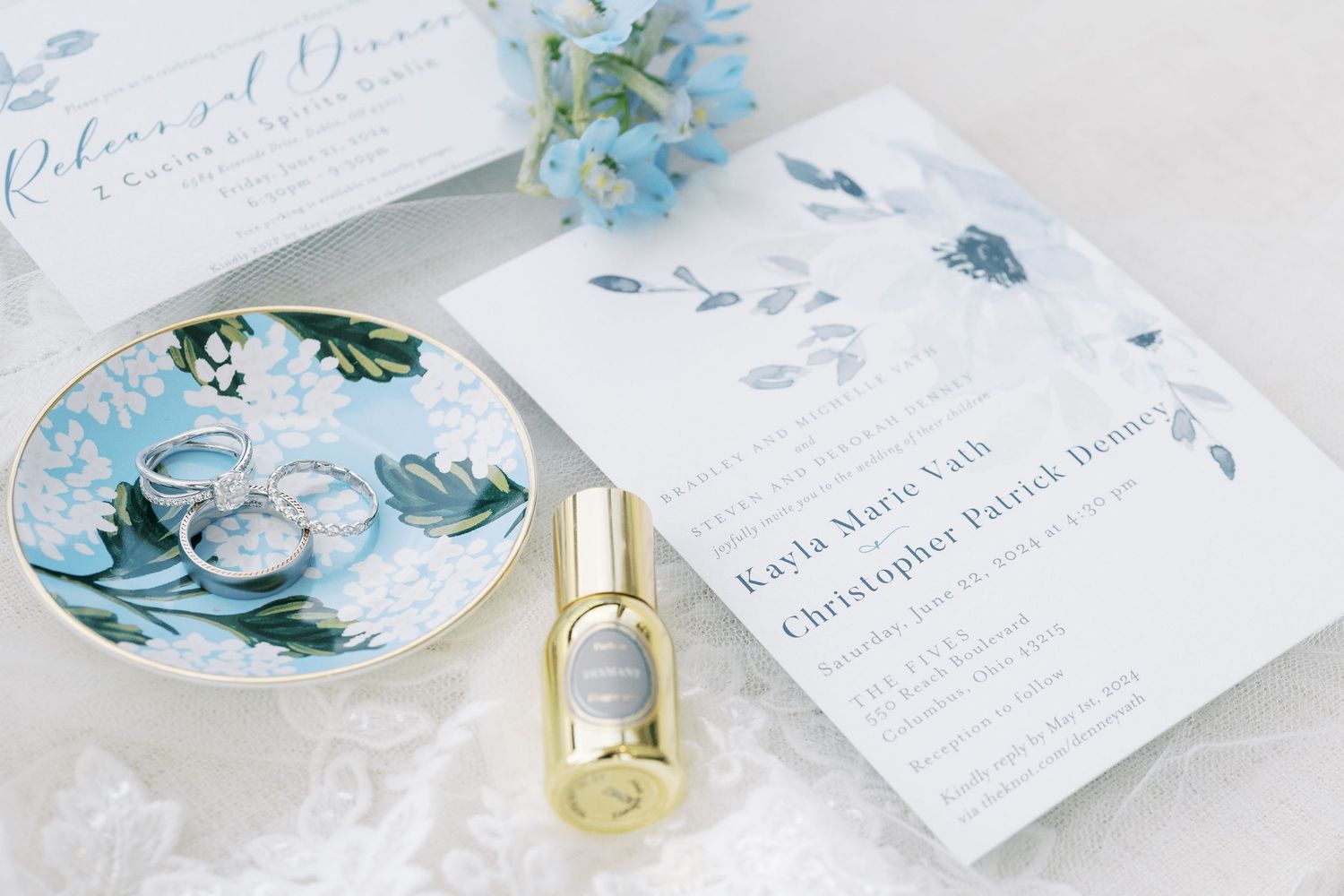 Wedding invitation suite in soft blue and white with floral designs and gold accents laid out on marble surface.