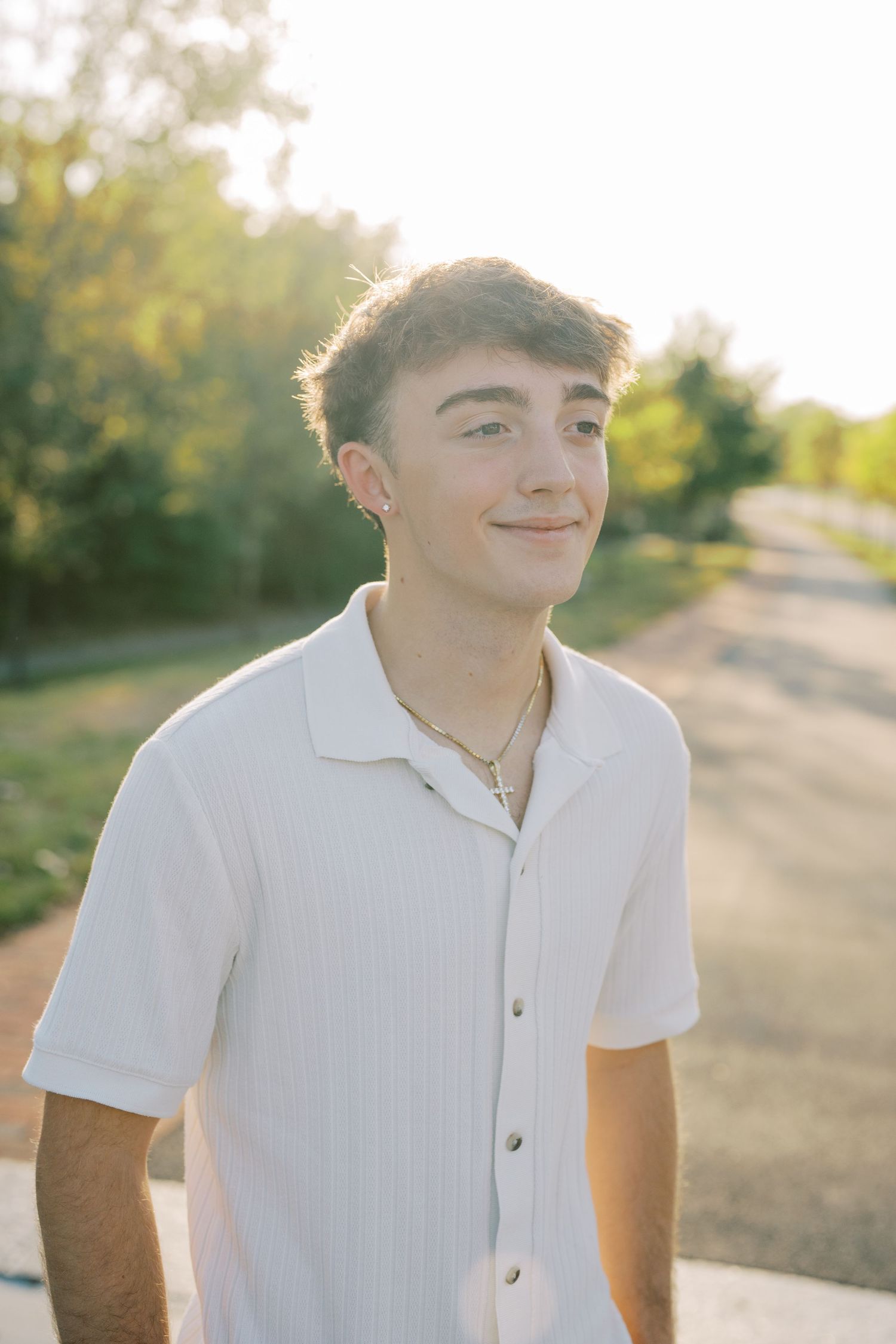 A person in a white polo shirt smiles warmly while standing outdoors in natural sunlight.