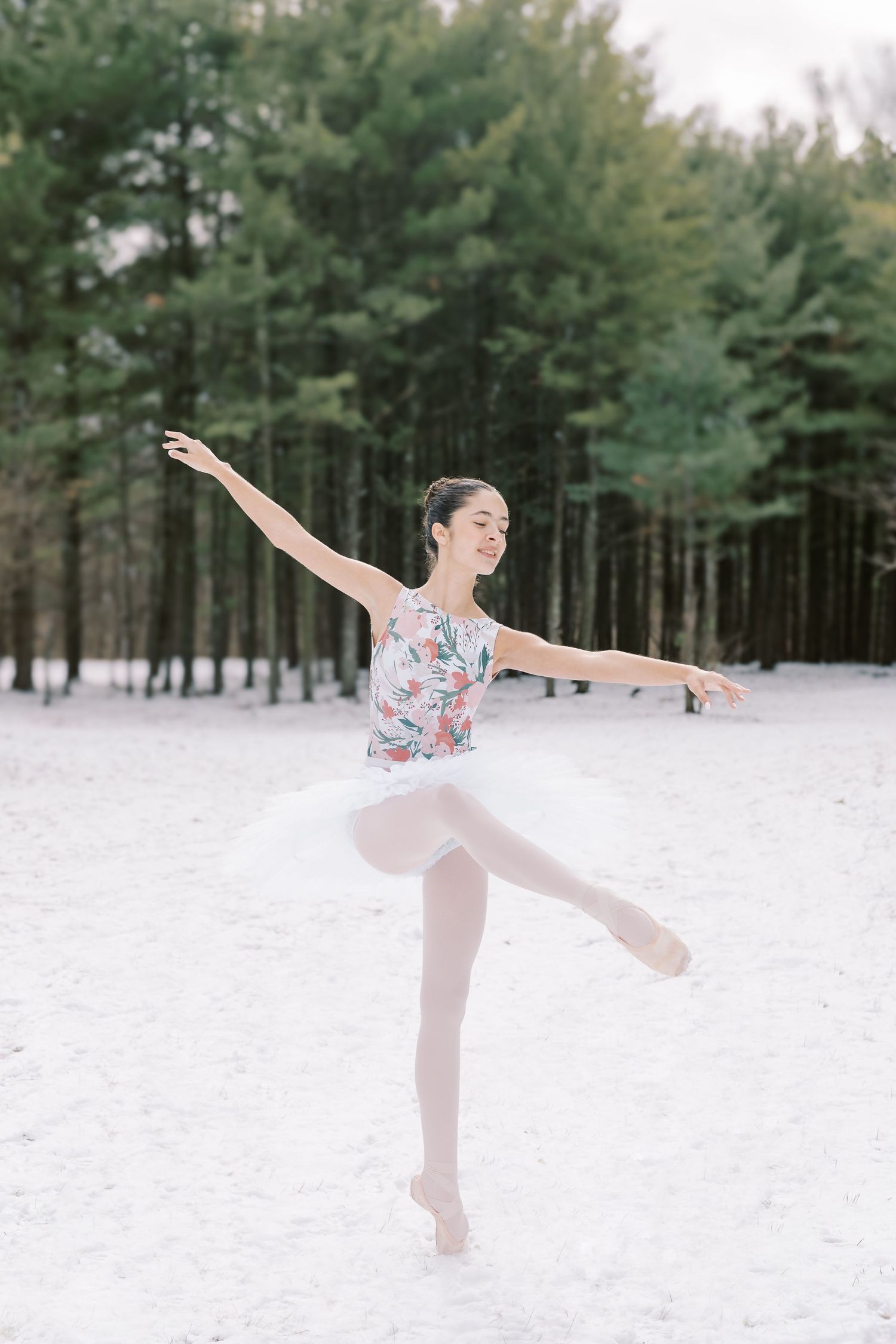 A ballet dancer performs graceful movements in the snow against a backdrop of evergreen trees in winter.