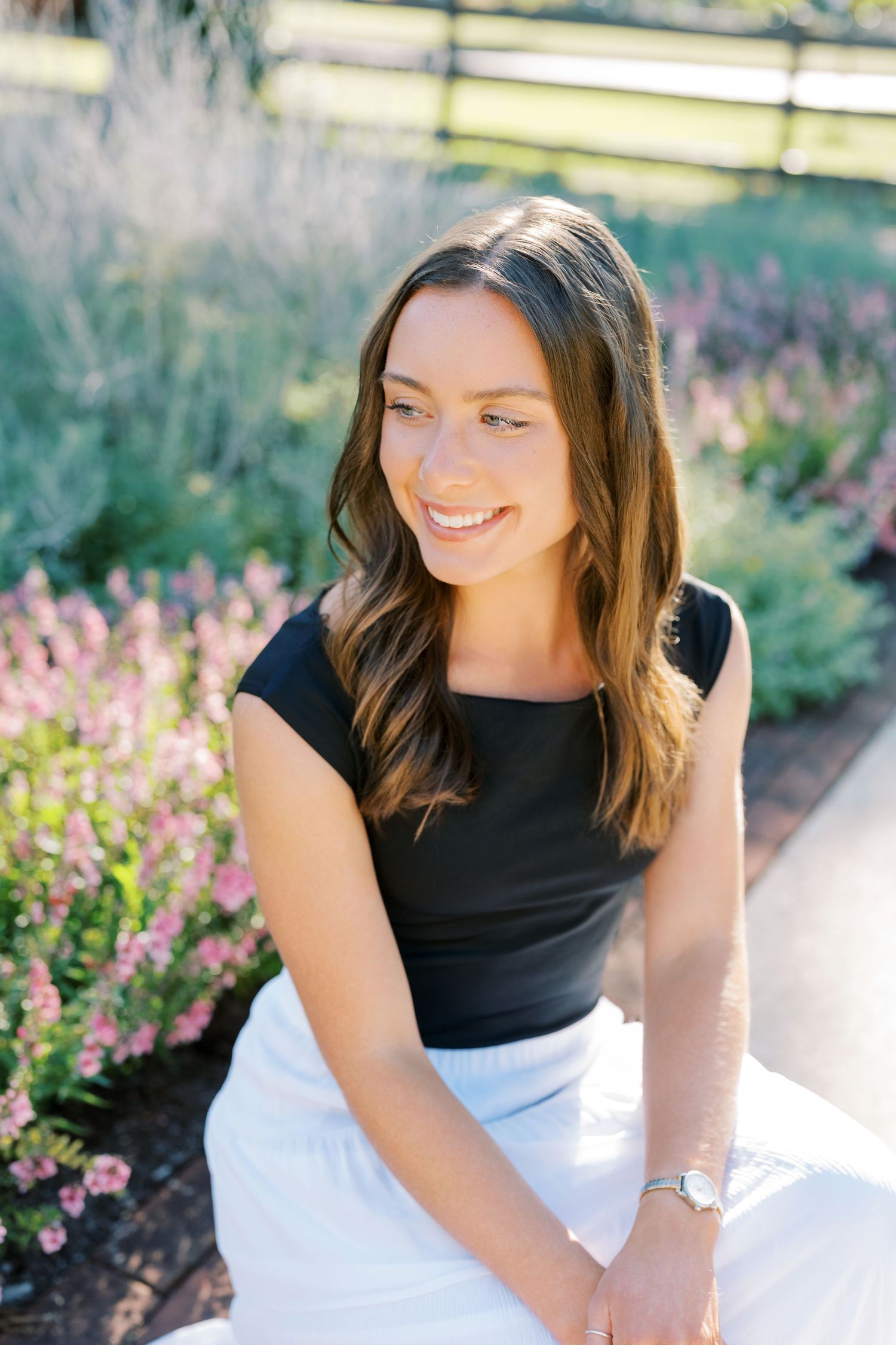 A person in a black and white dress sits outdoors smiling among blooming pink flowers in a garden setting.