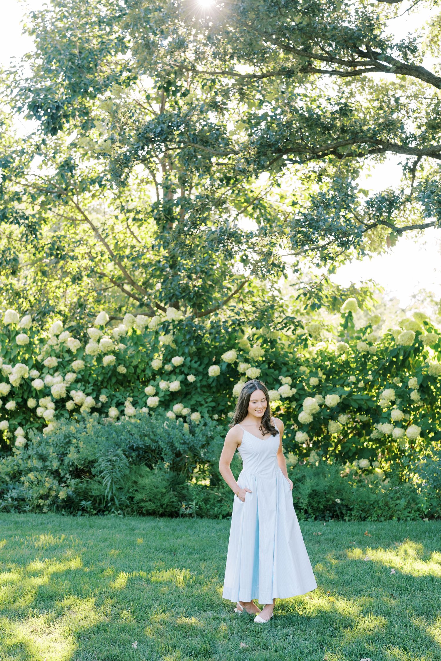 A person in a flowing white dress stands in a sunlit garden with blooming yellow flowers and lush greenery.