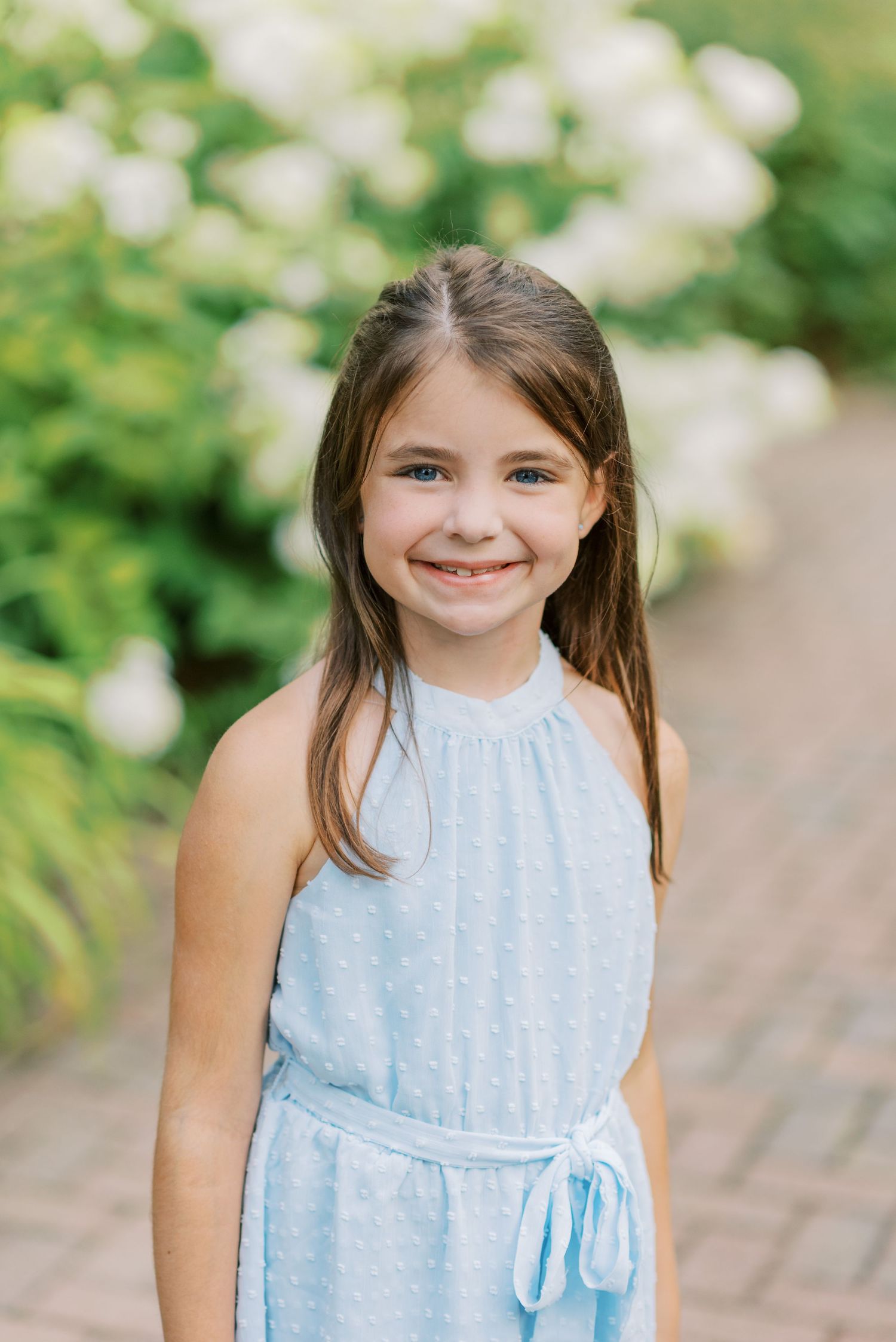 Young girl in light blue polka dot dress standing on brick path with white flowers in garden background.