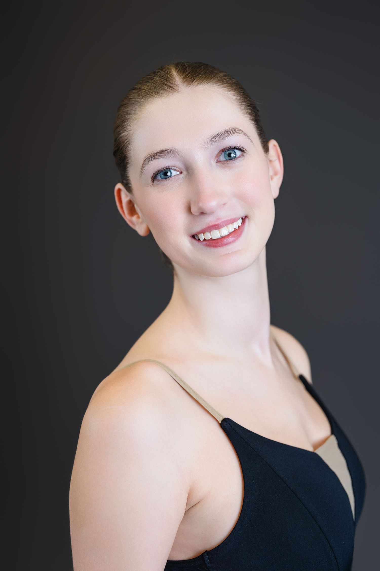 Portrait of a dancer in a black leotard smiling warmly against a dark background.