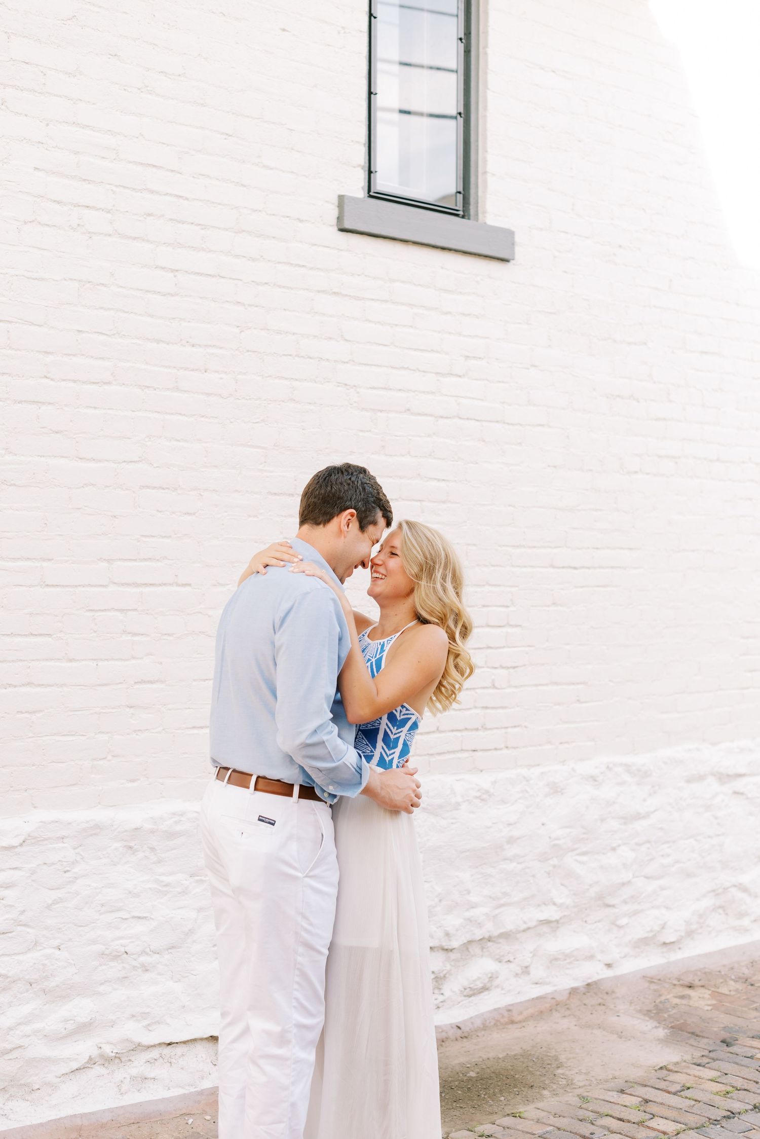 A romantic couple embraces against a white brick wall, wearing light summer attire in a tender moment.