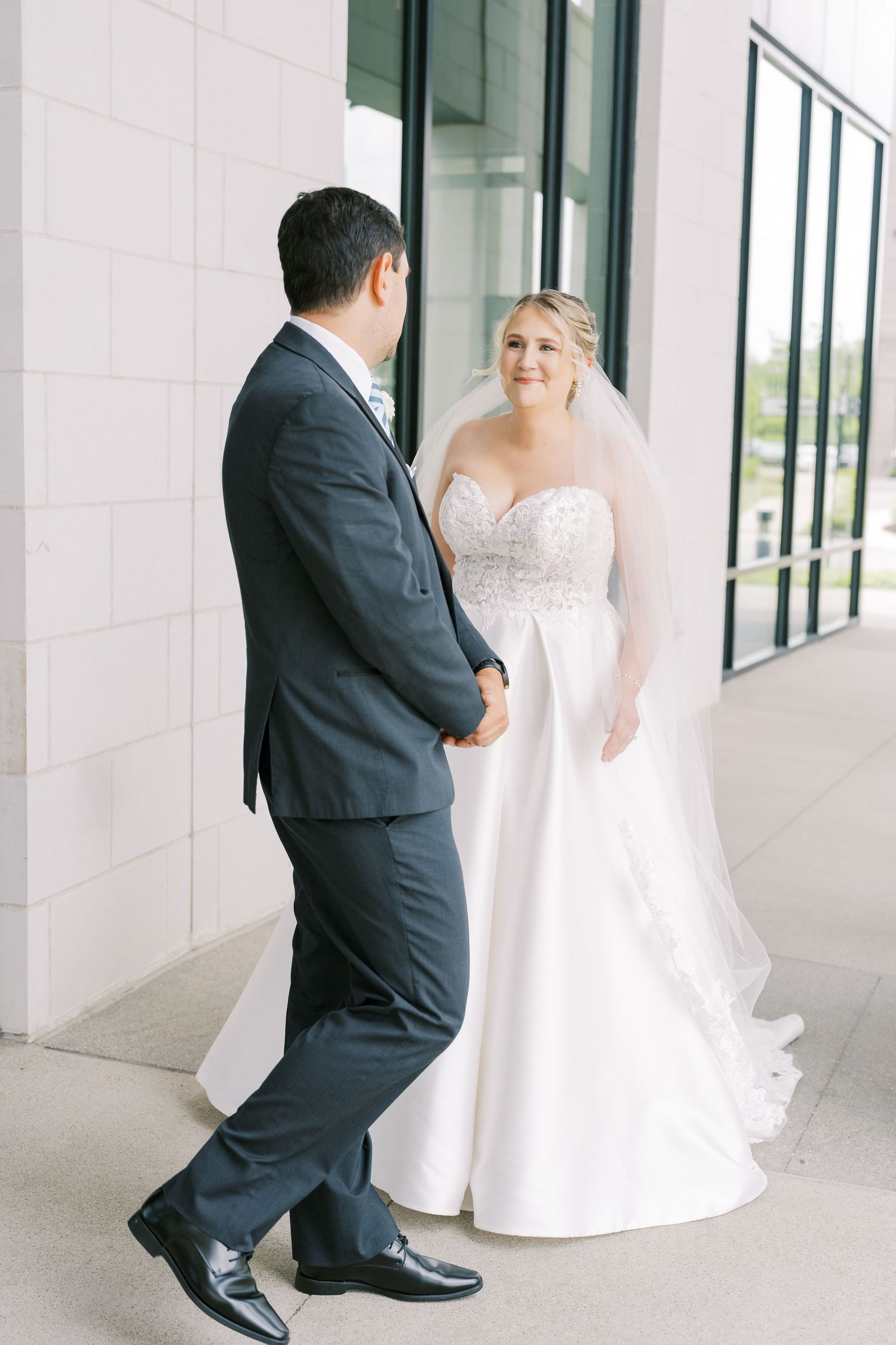 A bride in a white strapless gown and veil stands with a groom in a dark suit during their first look moment.