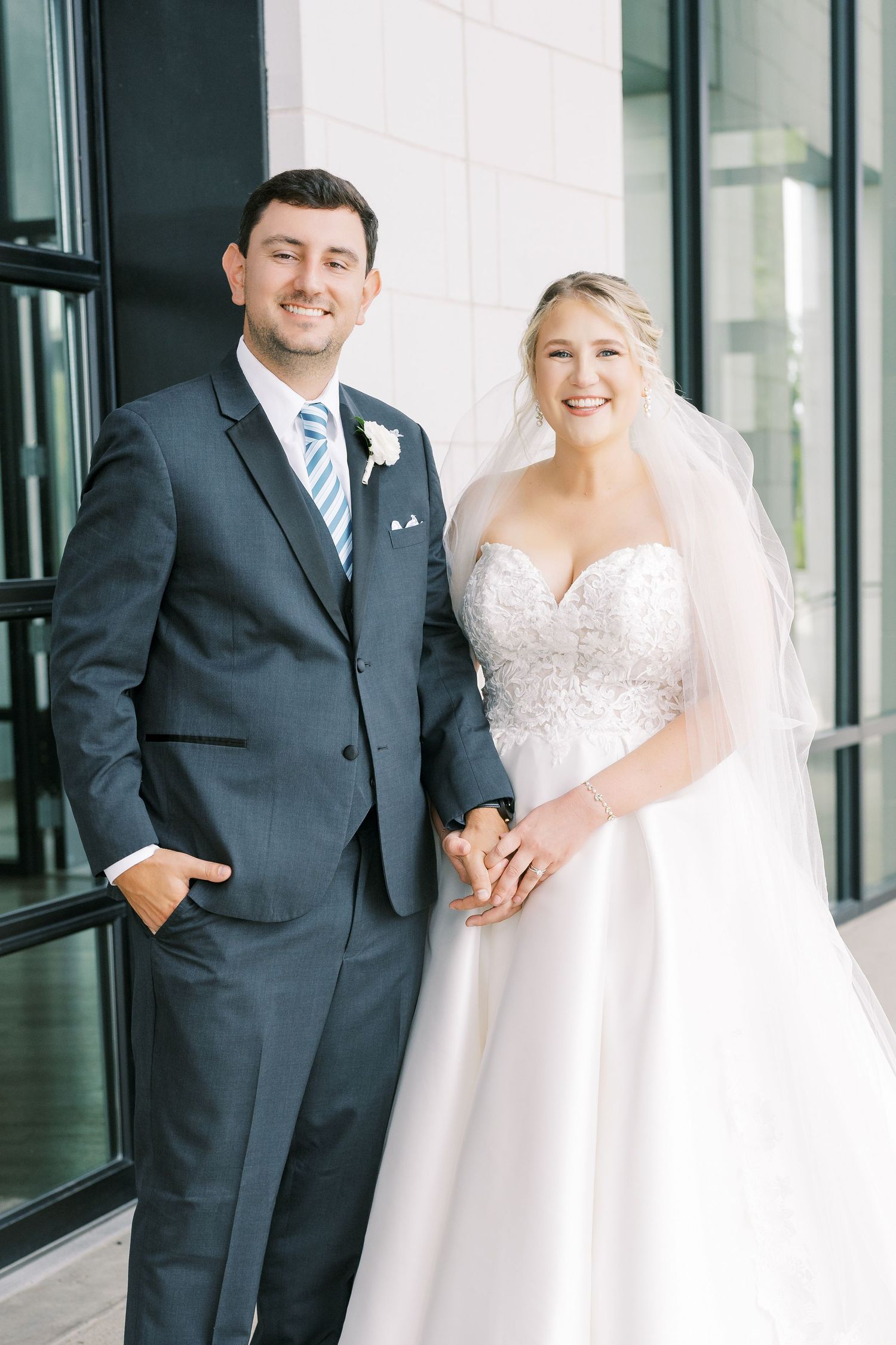 A couple shares joyful moments in their wedding attire against a modern glass building backdrop.