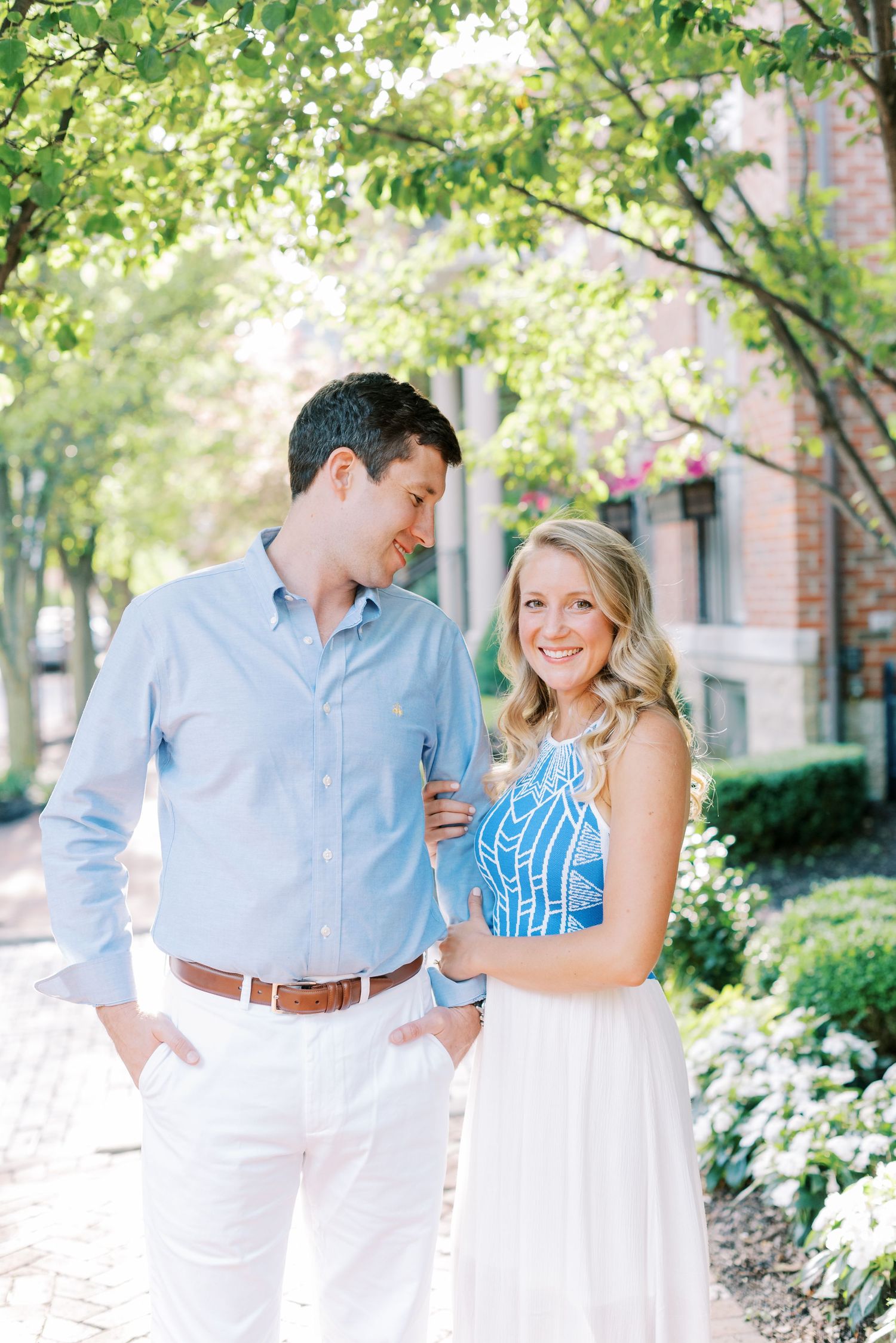 A couple in coordinated blue and white outfits pose together on a tree-lined path near a brick building.