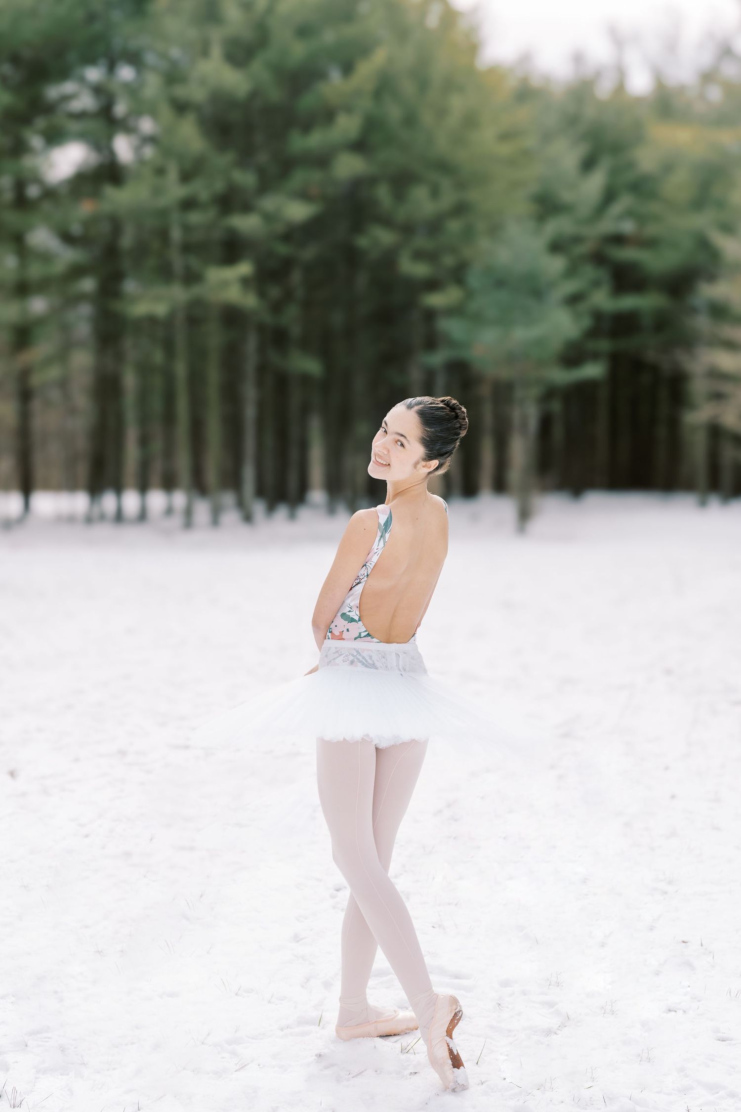 A dancer in white ballet attire performs graceful poses in a snowy forest setting with evergreen trees in the background.
