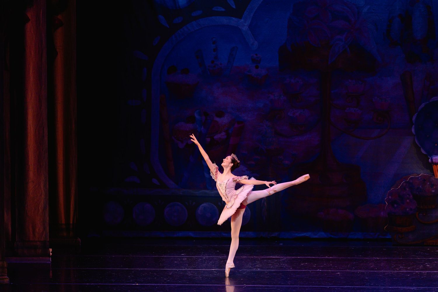 Ballet dancer in pink tutu performs graceful arabesque pose on dimly lit stage with blue backdrop.