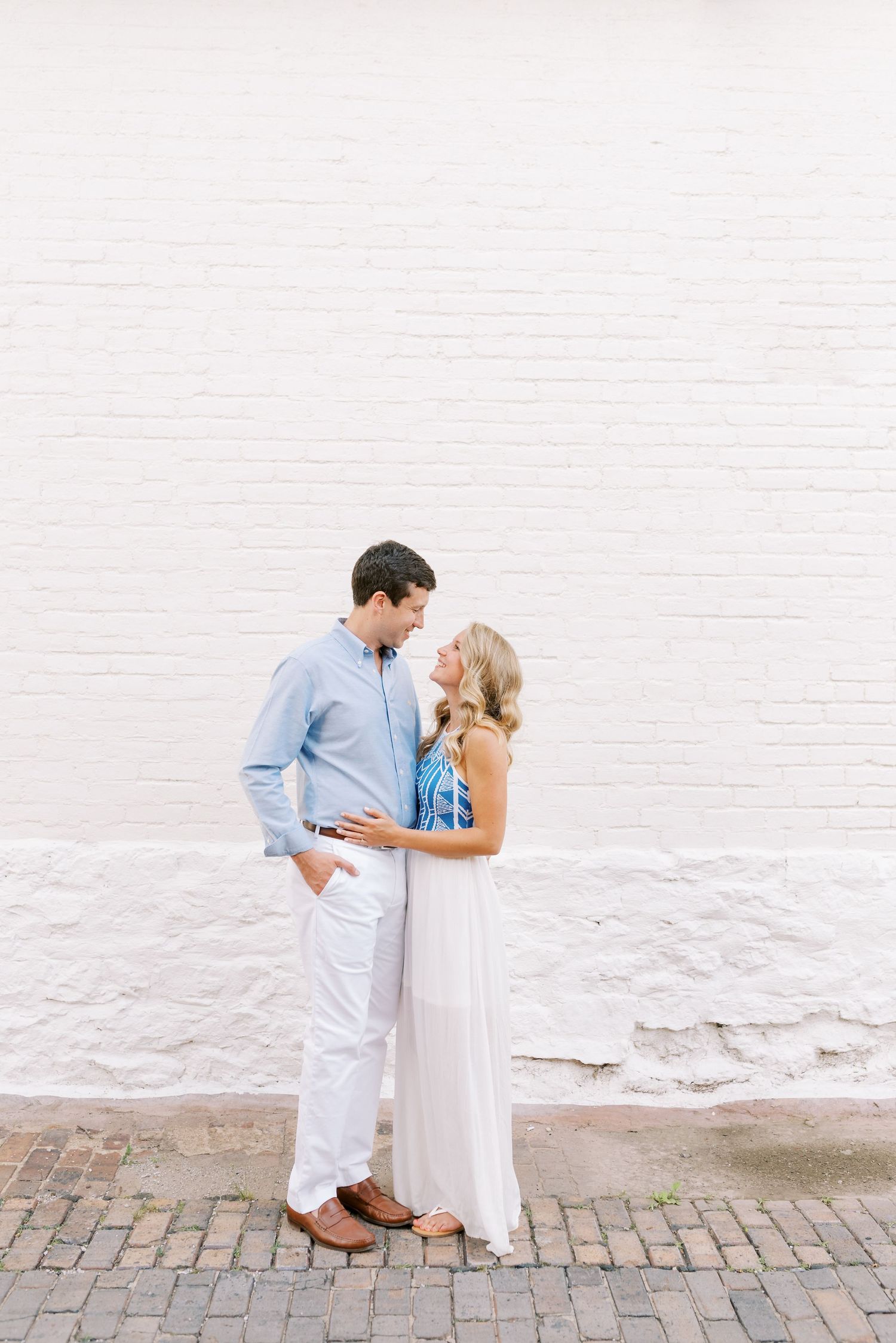 Romantic couple in light blue and white outfits pose against a white brick wall for engagement photos.