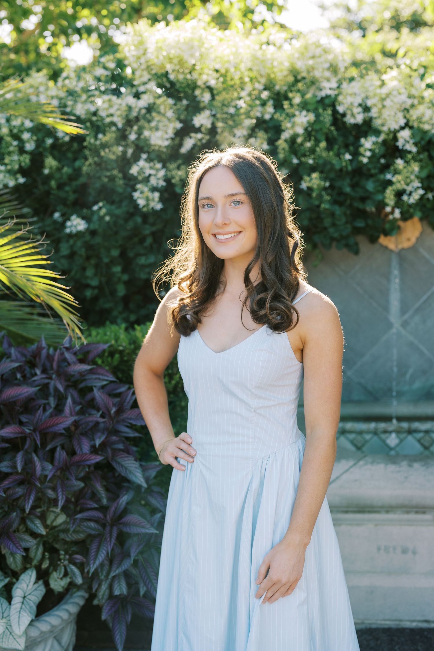 A light blue sleeveless dress is worn in a garden setting with flowering white bushes and tropical plants in the background.