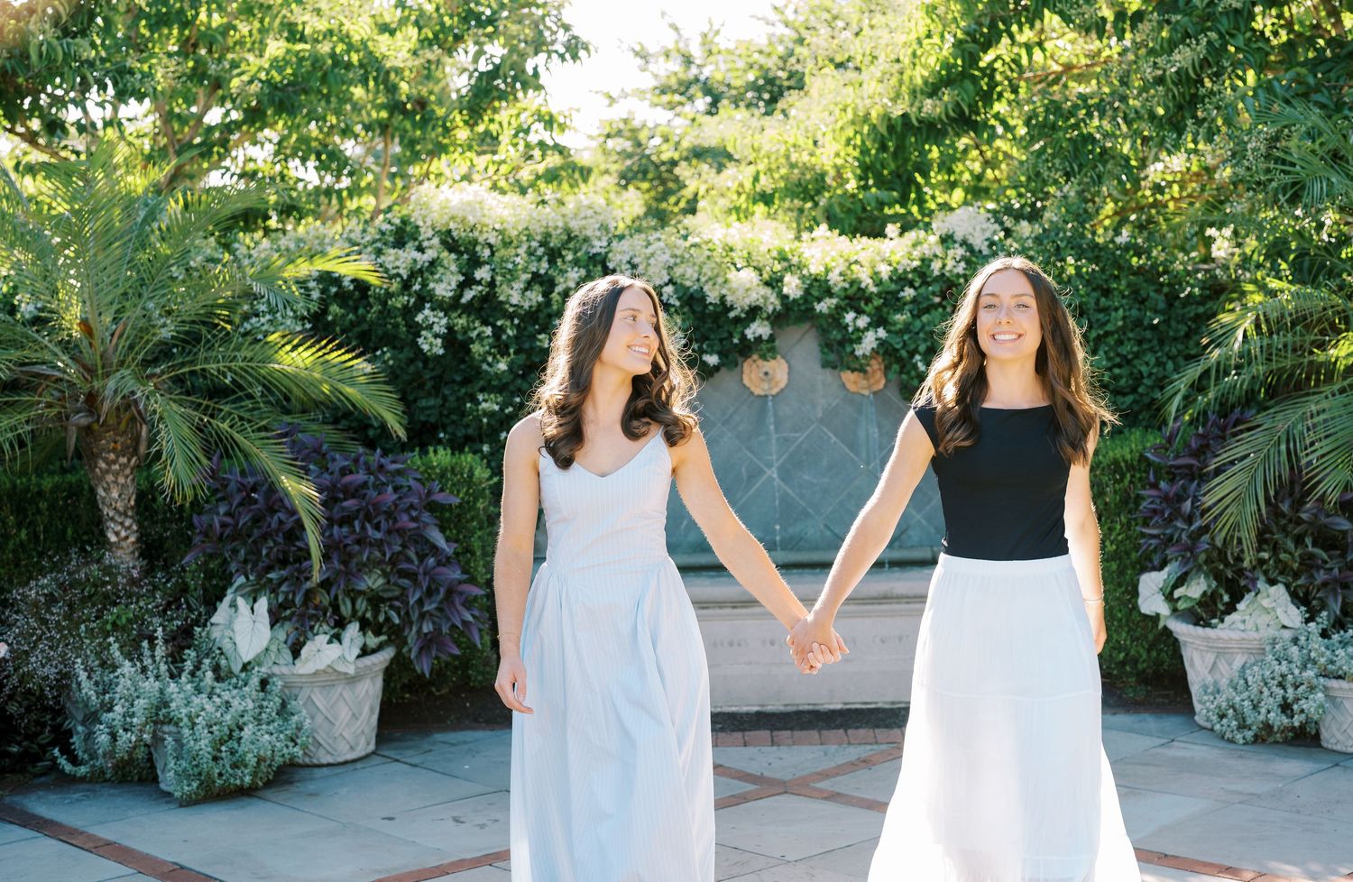 Two people in white dresses walk hand in hand through a lush garden setting on a sunny day.