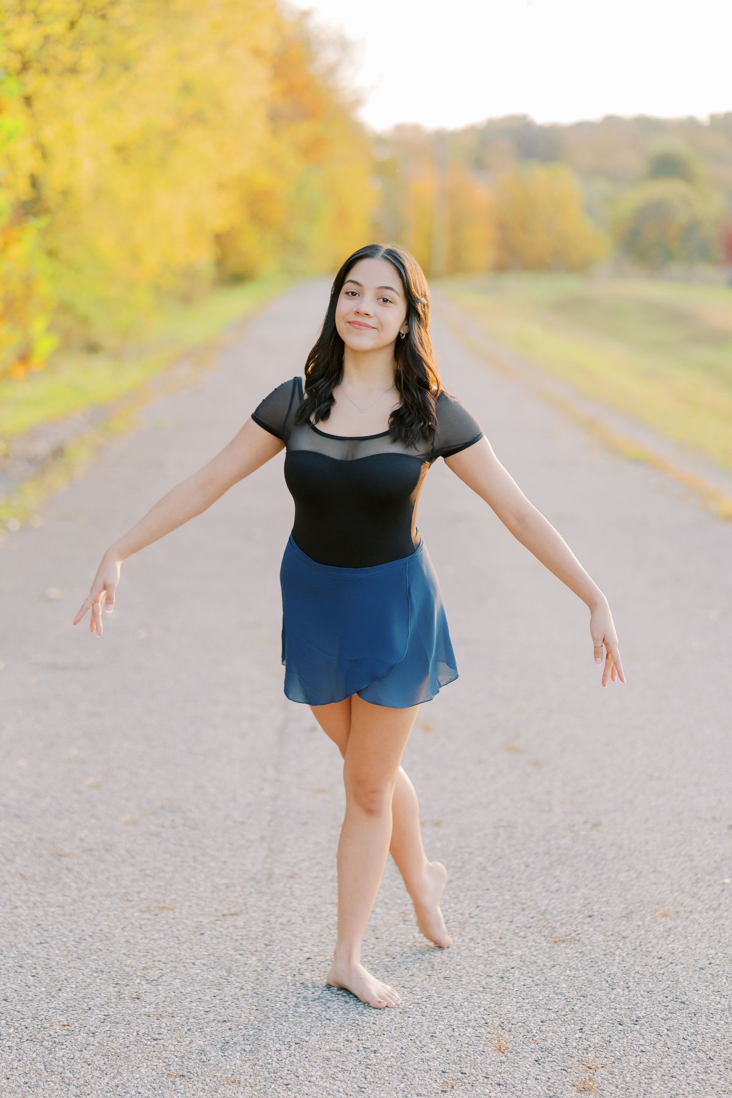 A figure in a black top and blue shorts dances gracefully on a gravel path lined with autumn trees.