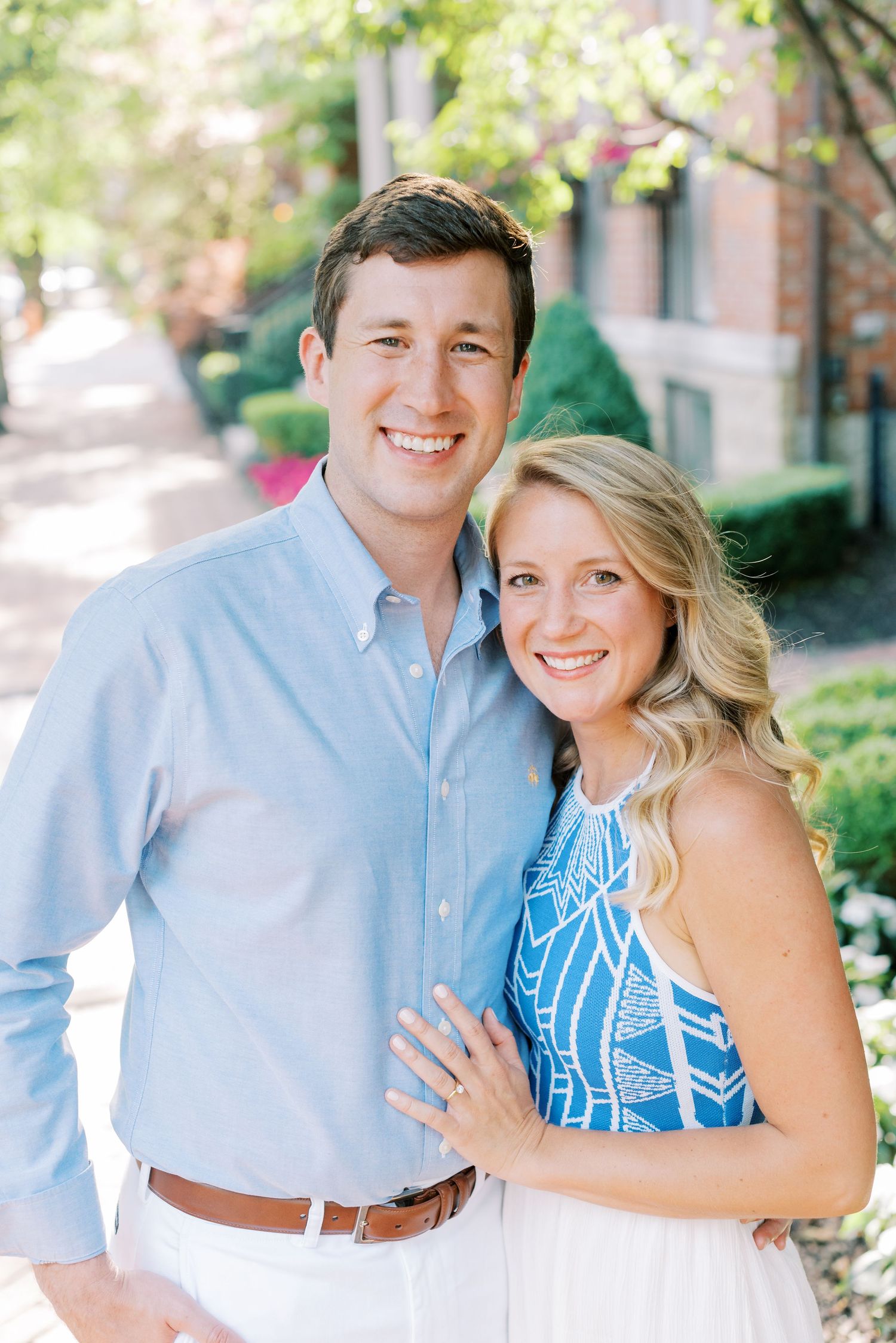 A couple poses together outdoors in spring, wearing a light blue button-down shirt and blue patterned sleeveless dress.