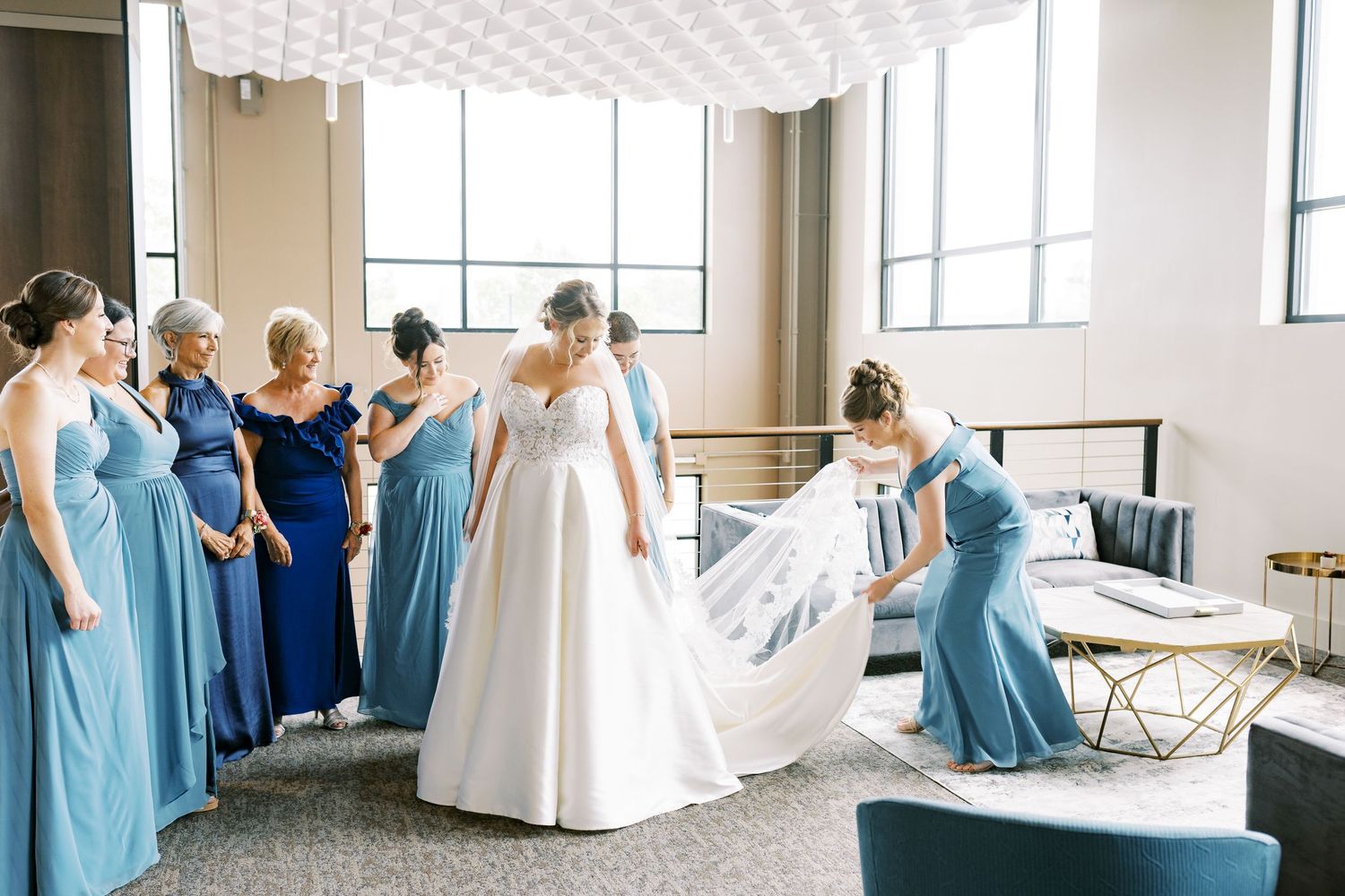 Bridesmaids in light blue dresses gather in a sunlit room during wedding preparations.