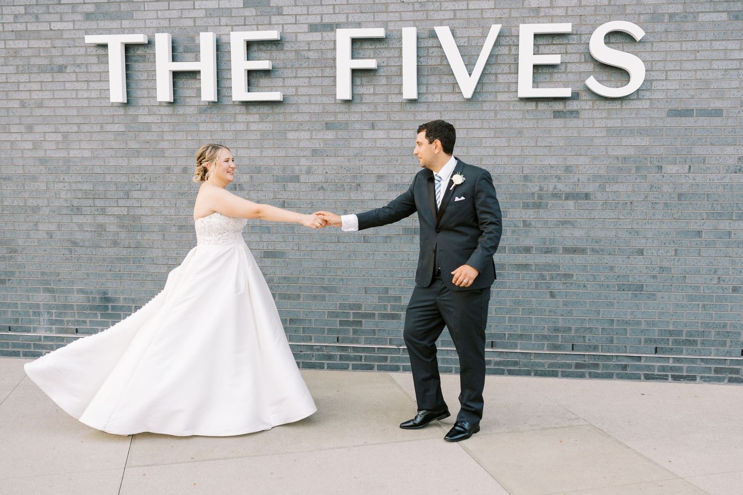 A couple in wedding attire dancing playfully in front of a gray brick wall with 'THE FIVES' signage.