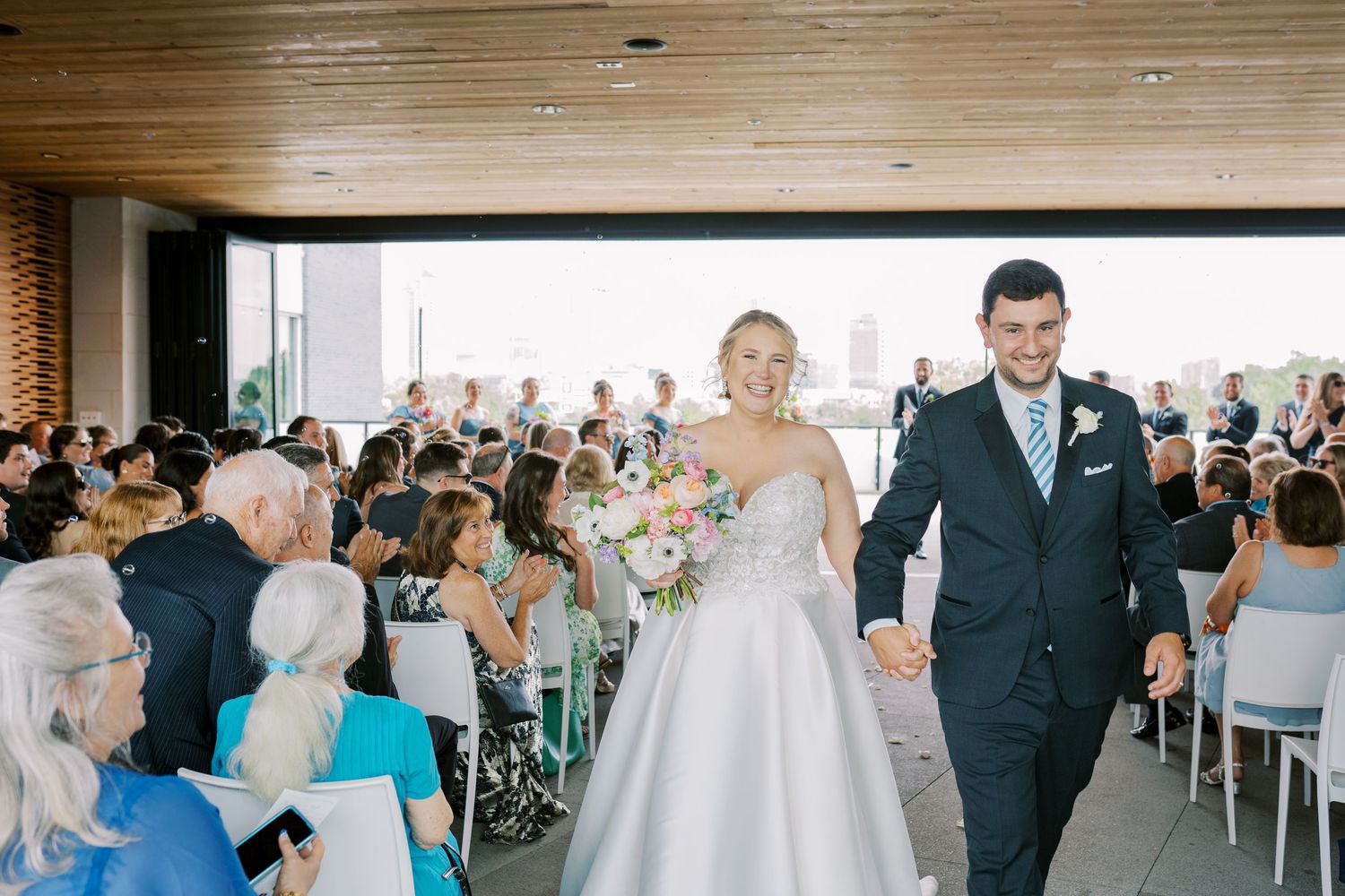 Newlyweds walk down the aisle together at an indoor wedding ceremony while guests look on from their seats.