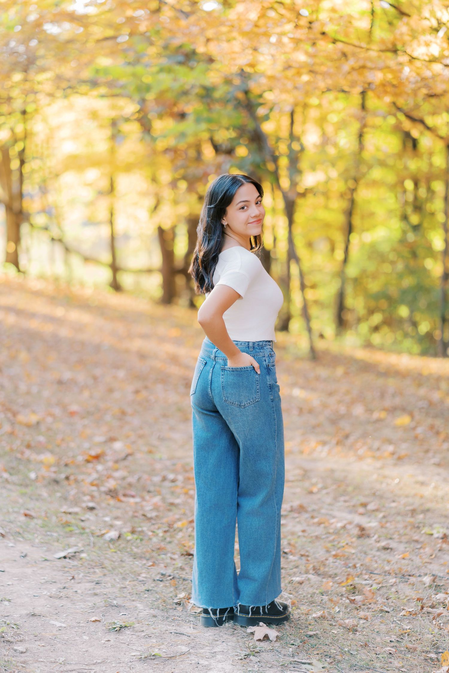A relaxed outdoor portrait showing a white t-shirt and wide-leg denim jeans against a golden autumn forest backdrop.