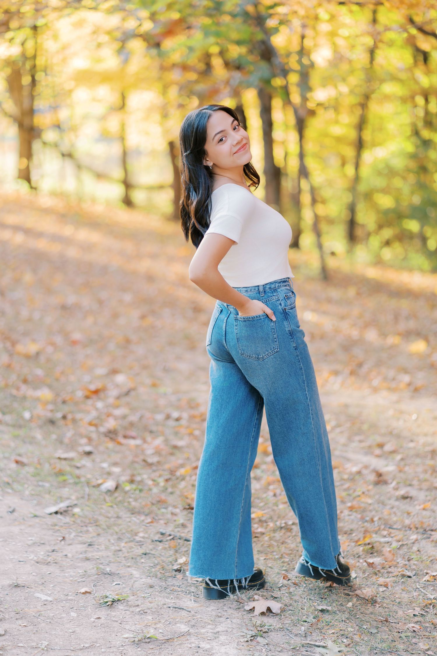 A person in a white top and wide-leg jeans poses on a forest path surrounded by golden autumn foliage.