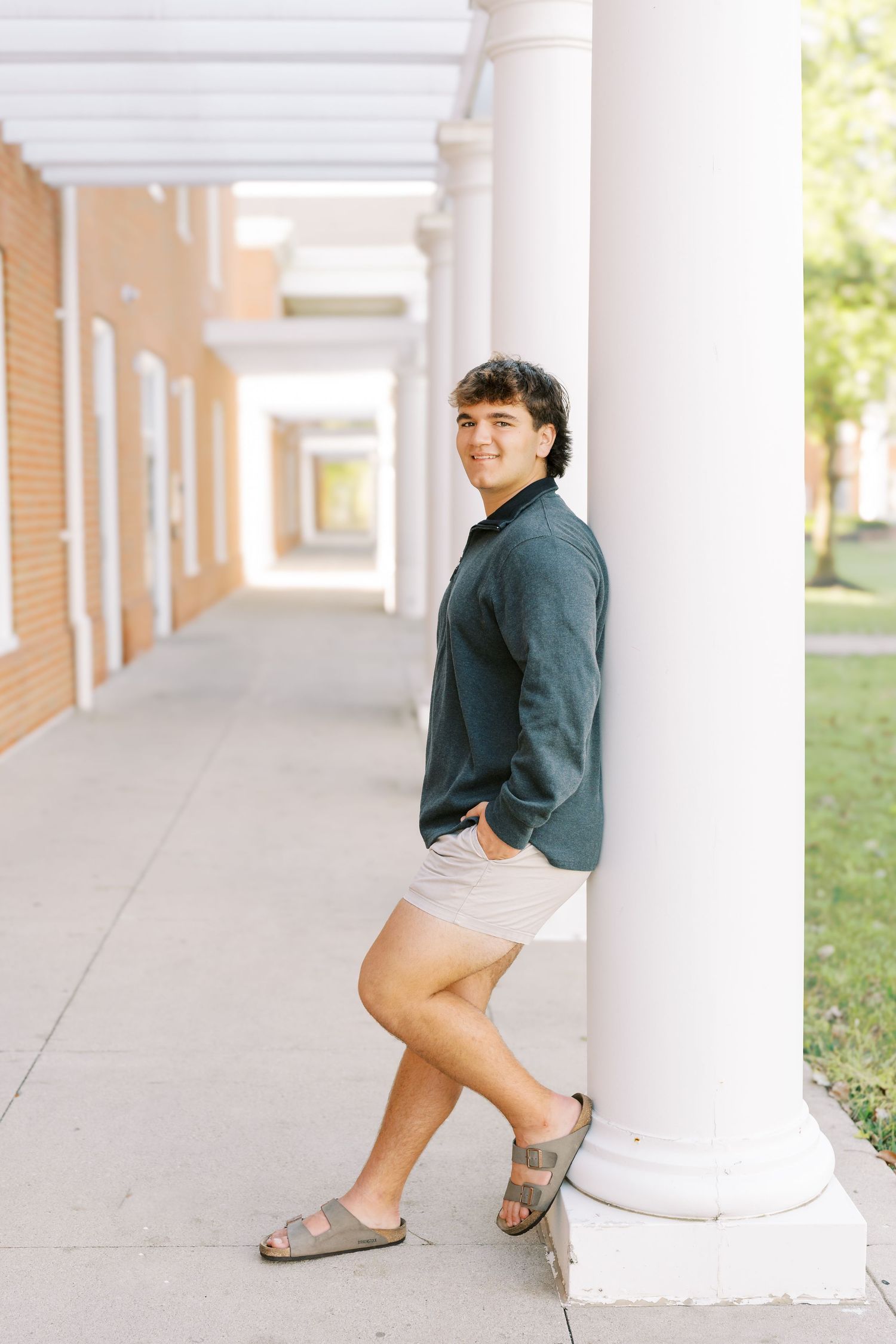 A person in a dark sweater and beige shorts poses casually against white columns in an outdoor walkway.
