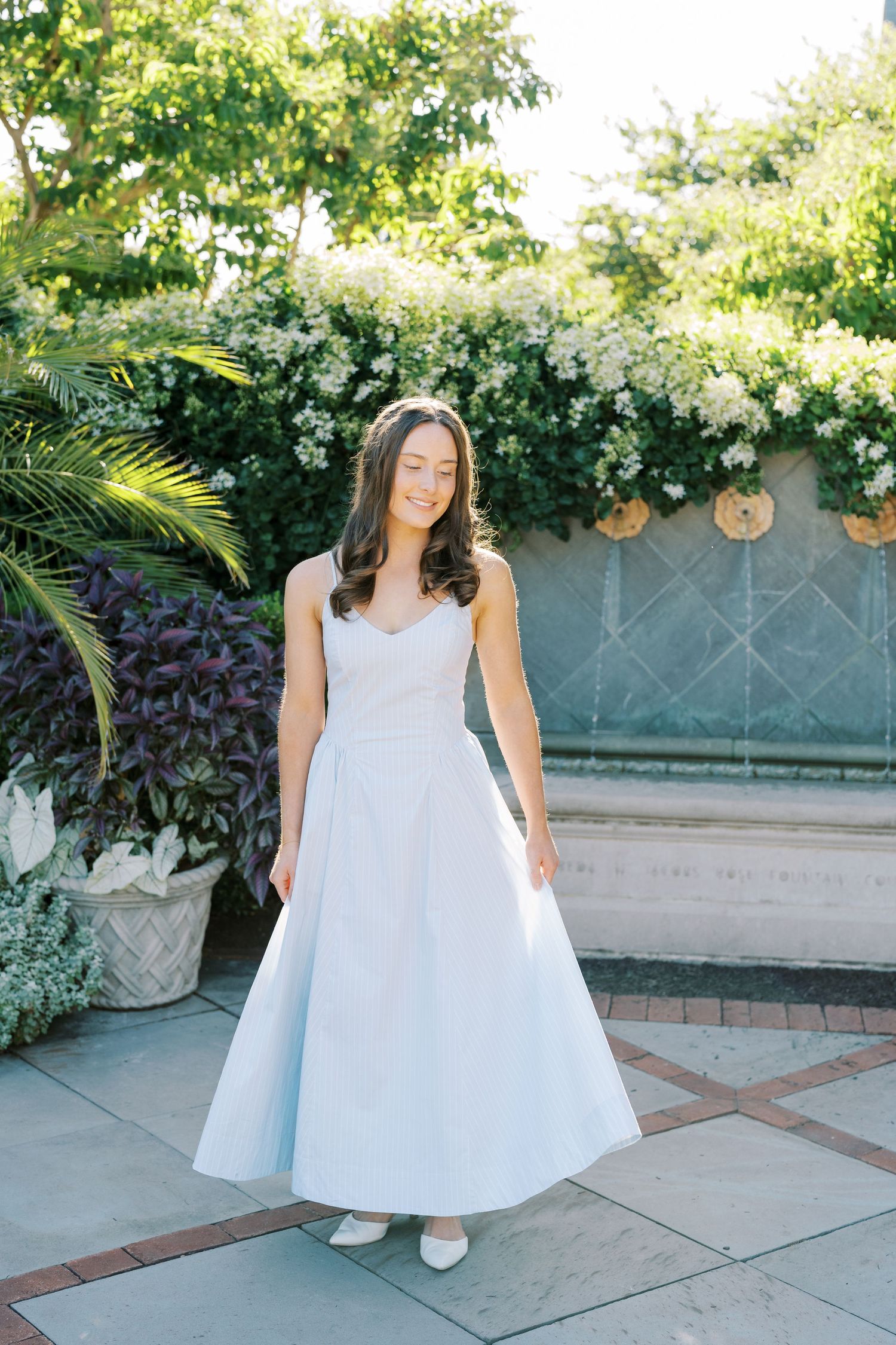 A person in a flowing white dress twirls on a paved patio surrounded by lush greenery and hanging lanterns.