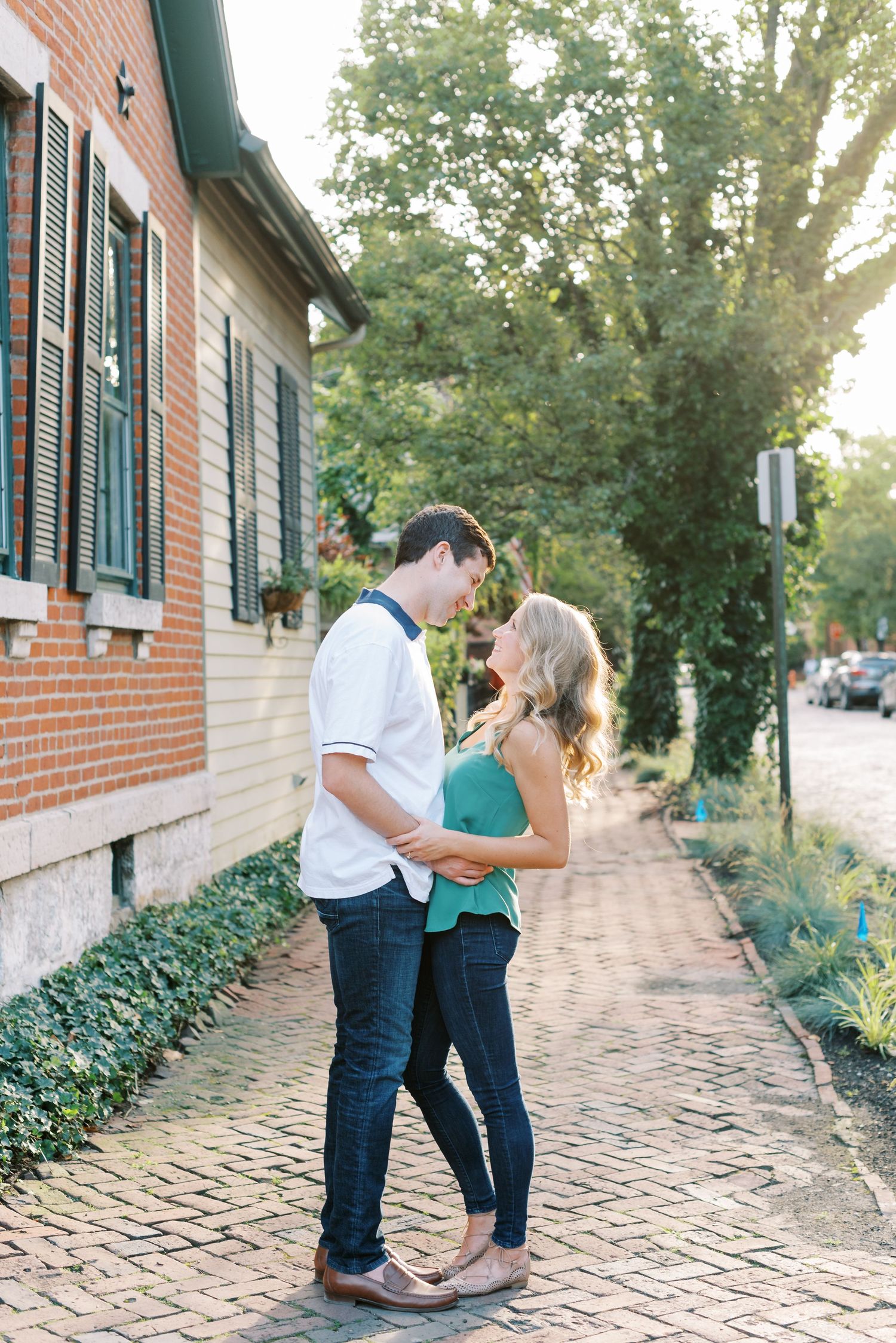 A couple shares a romantic moment on a brick sidewalk outside a historic building with green shutters.