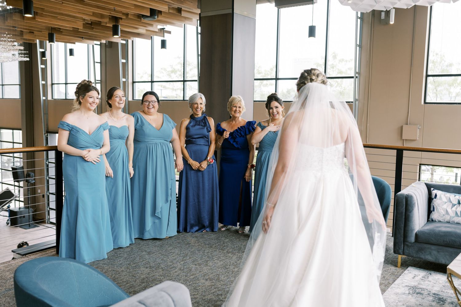 Bridesmaids in blue dresses help prepare the bride in a sunlit room with floor-to-ceiling windows.
