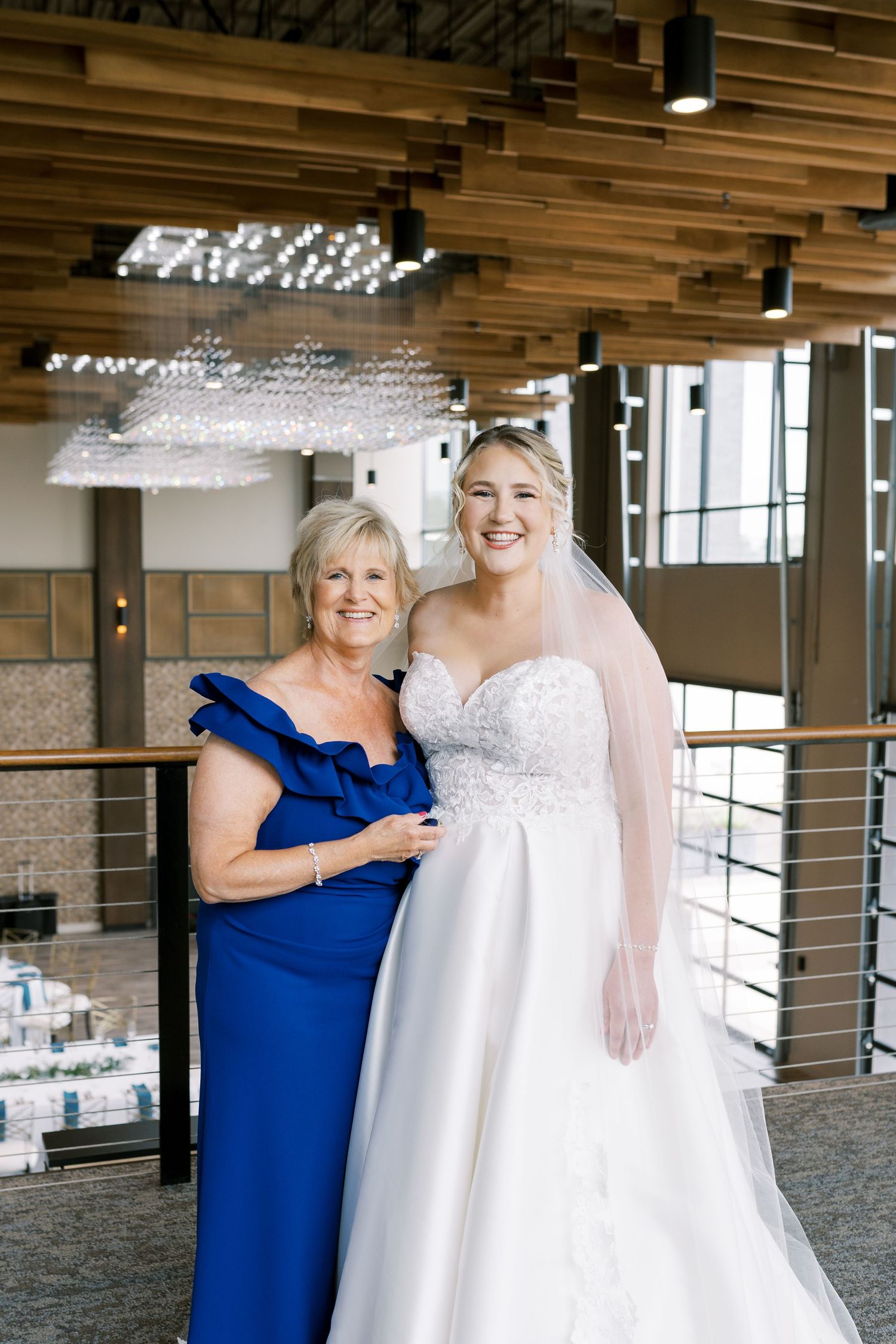 A bride in a white lace wedding dress shares a joyful moment with someone wearing royal blue in an elegant venue.