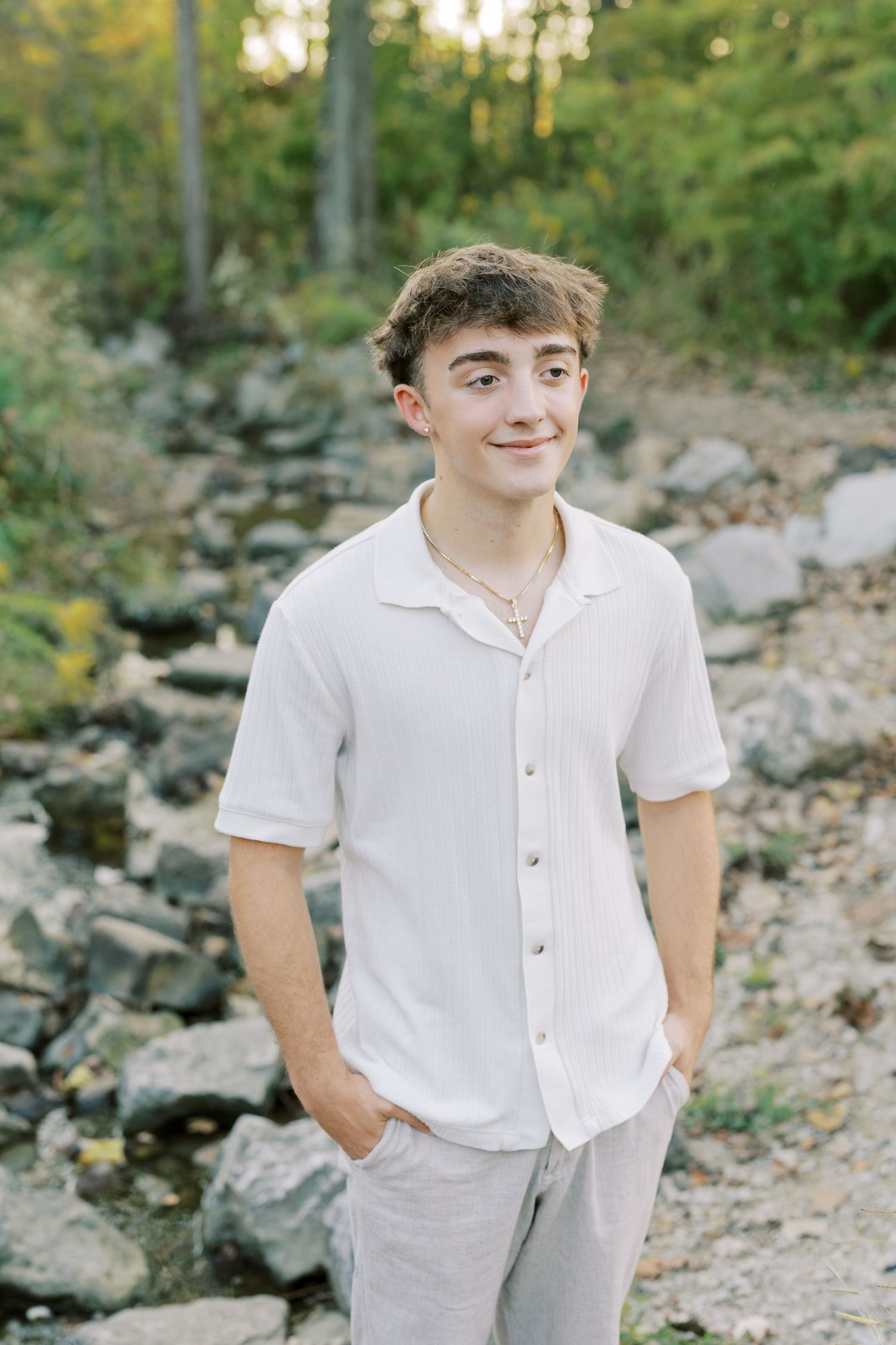 A person in a white button-down shirt and light pants poses casually on a rocky outdoor path during a sunny day.