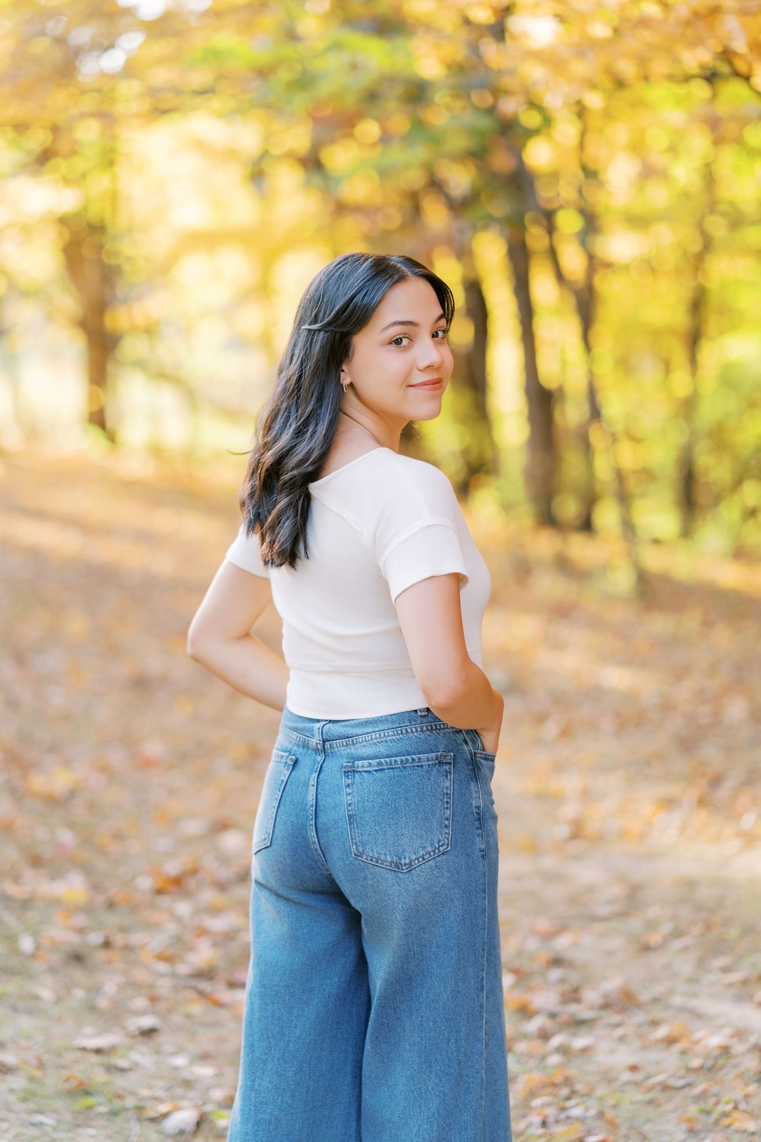 A casual autumn fashion photo showing someone in a white t-shirt and wide-leg jeans standing on a leaf-covered path.