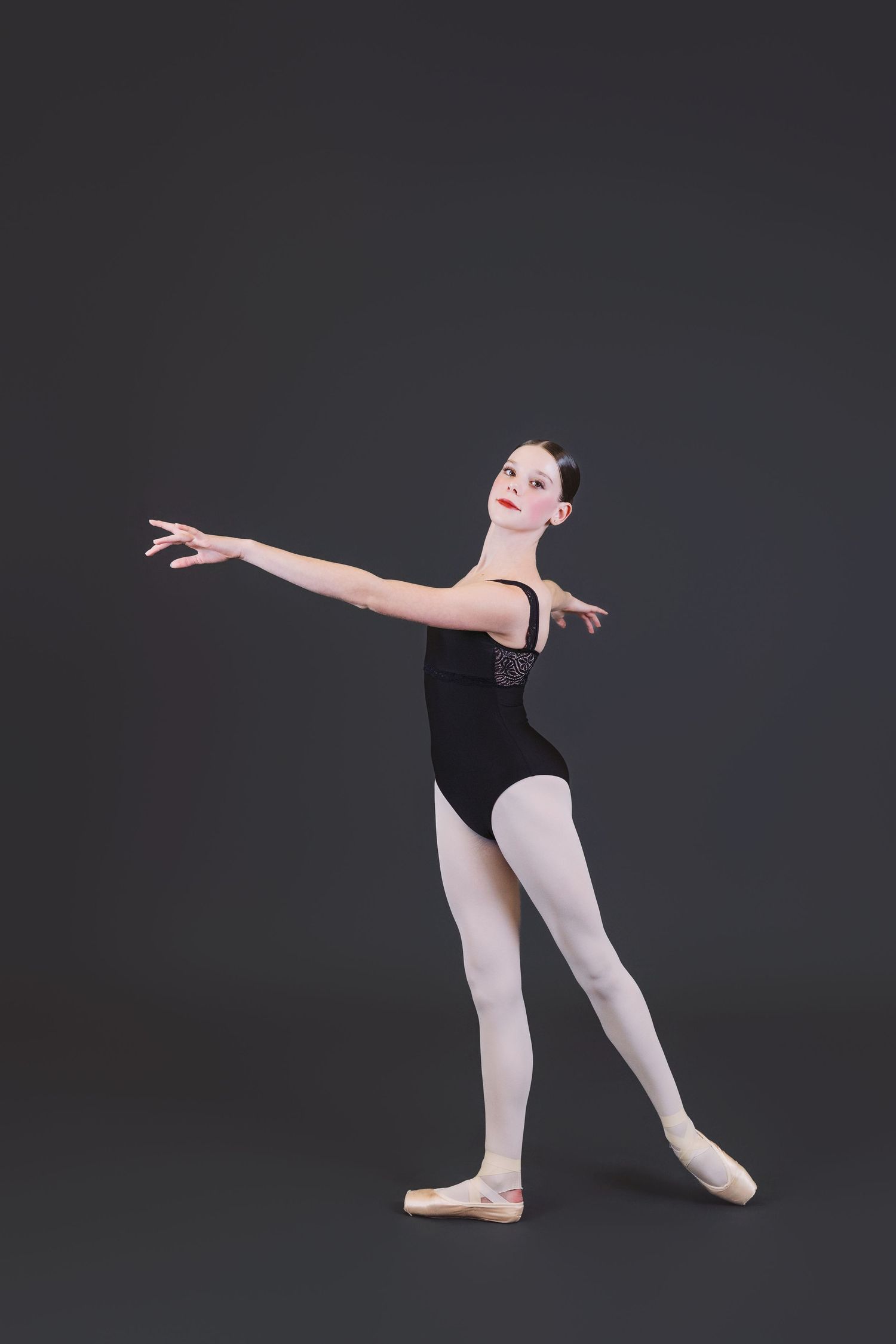 A ballet dancer in black leotard and white tights performs an arabesque pose against a dark studio background.