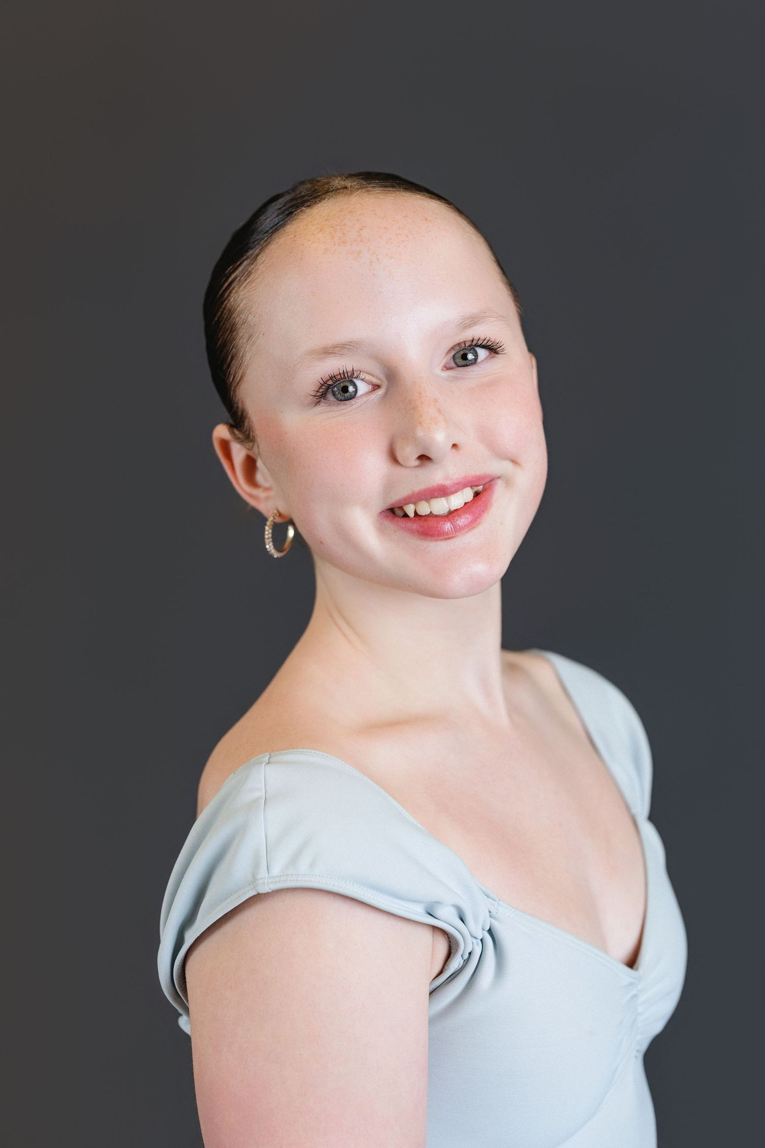 A dancer in a light blue leotard poses against a gray background with a bright smile and small hoop earrings.