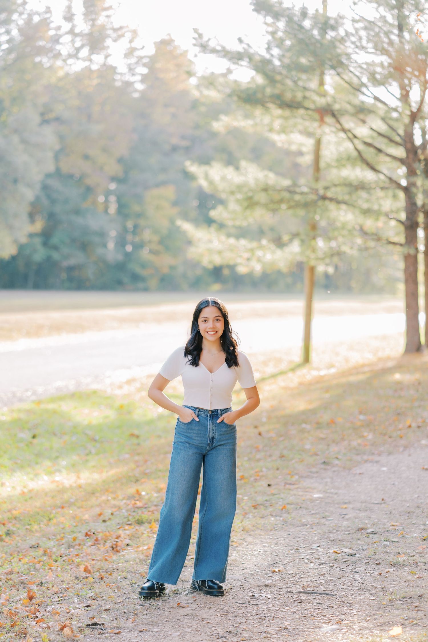 A person in a white top and wide-leg jeans stands on a path surrounded by autumn trees and soft sunlight.
