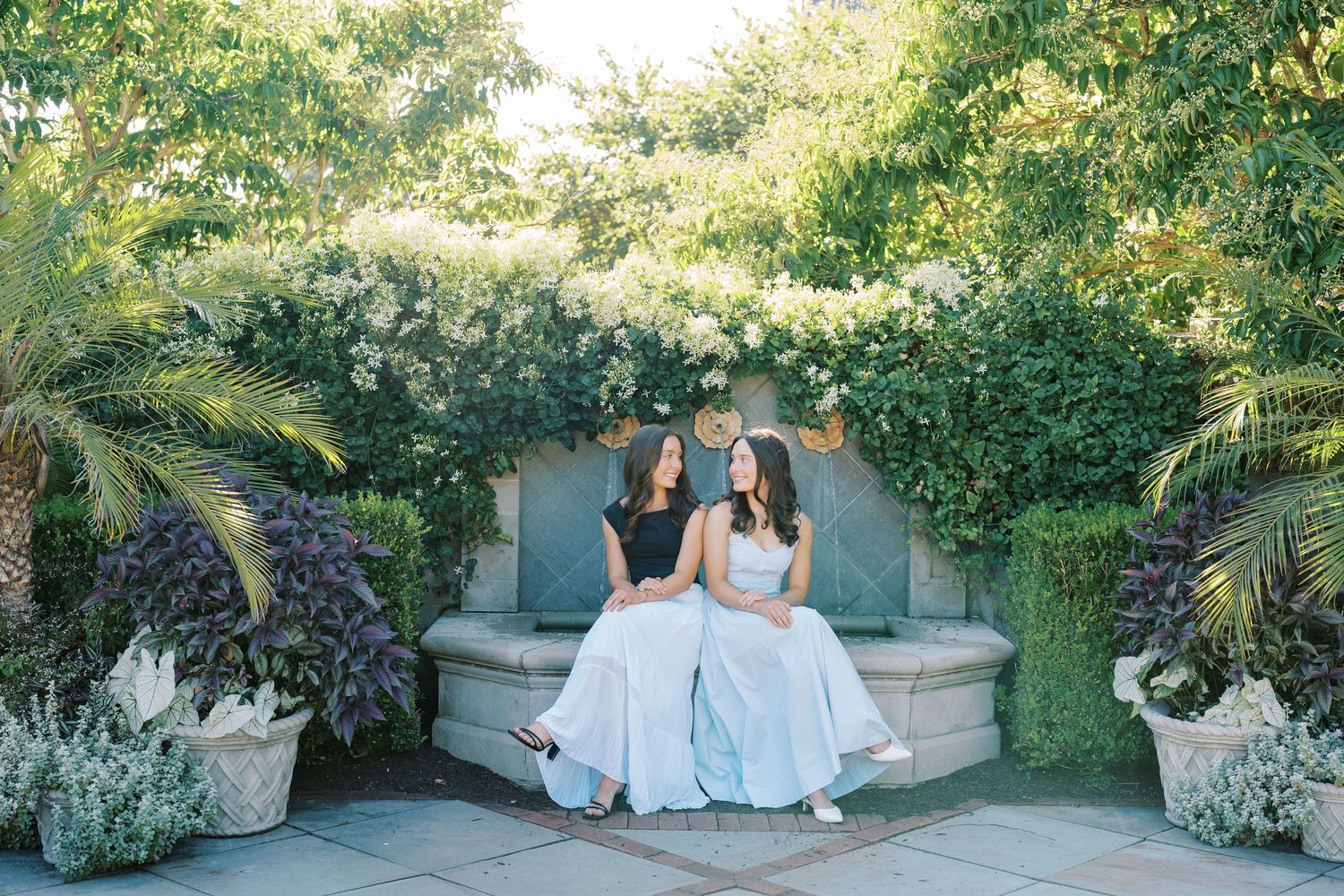 Two people in wedding attire sit together on a garden bench surrounded by lush greenery and flowers.