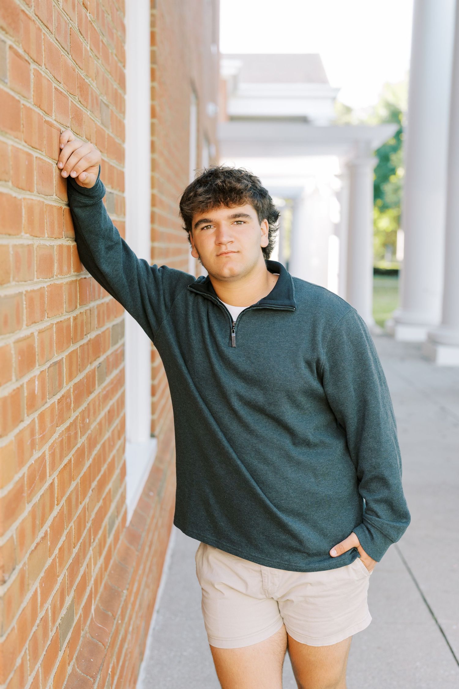 A person in a dark quarter-zip pullover and khaki shorts leans against a brick wall along a covered walkway.