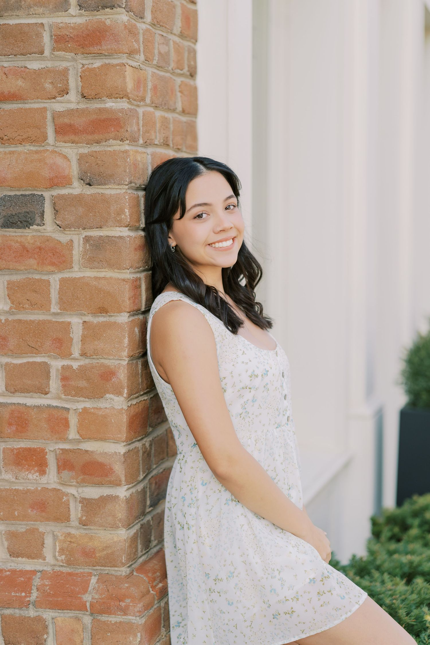 A person in a white lace dress poses against a brick wall near greenery in a soft natural light setting.