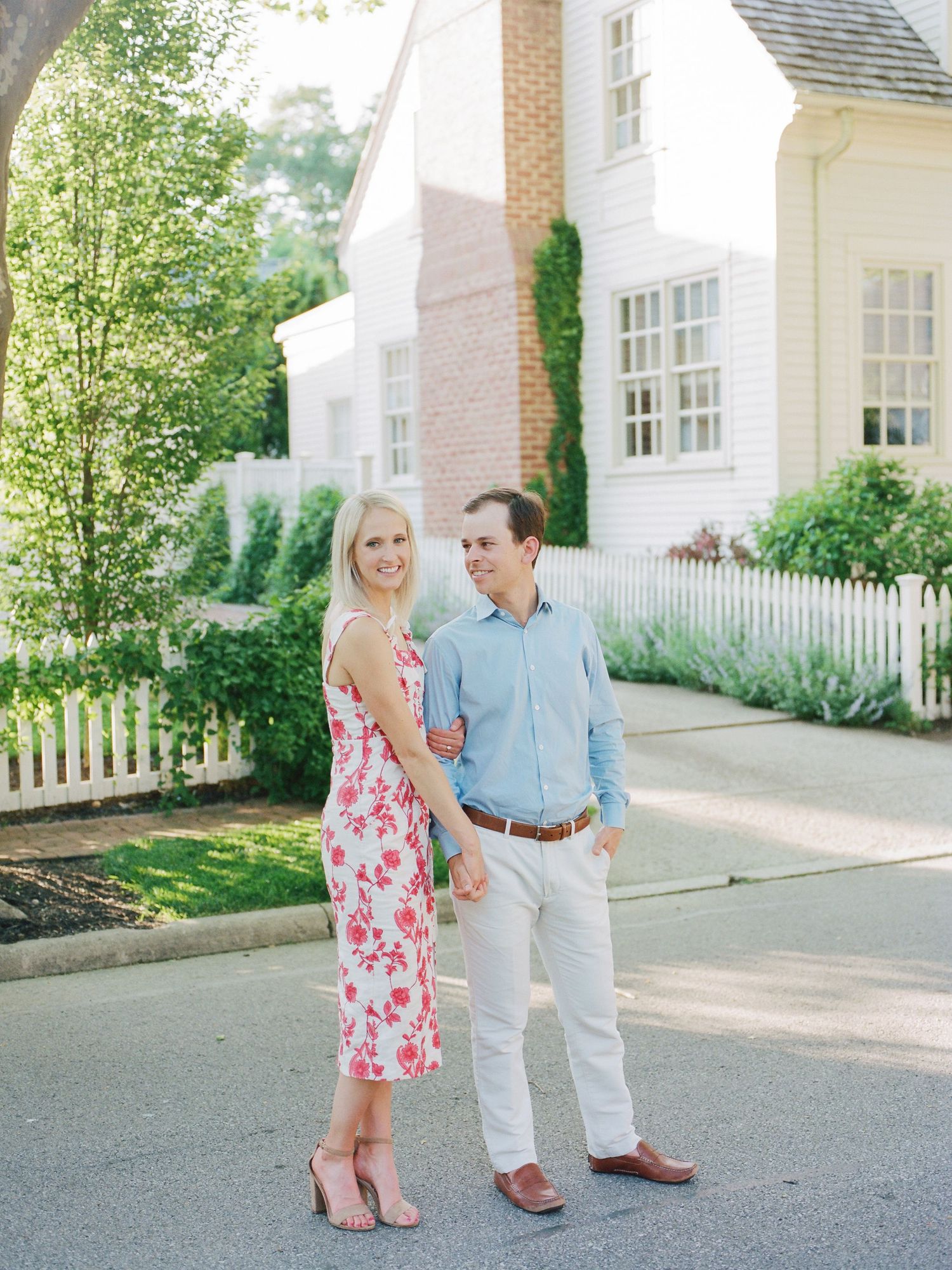 A couple shares a tender moment while walking together on a paved path beside a white house with a picket fence.