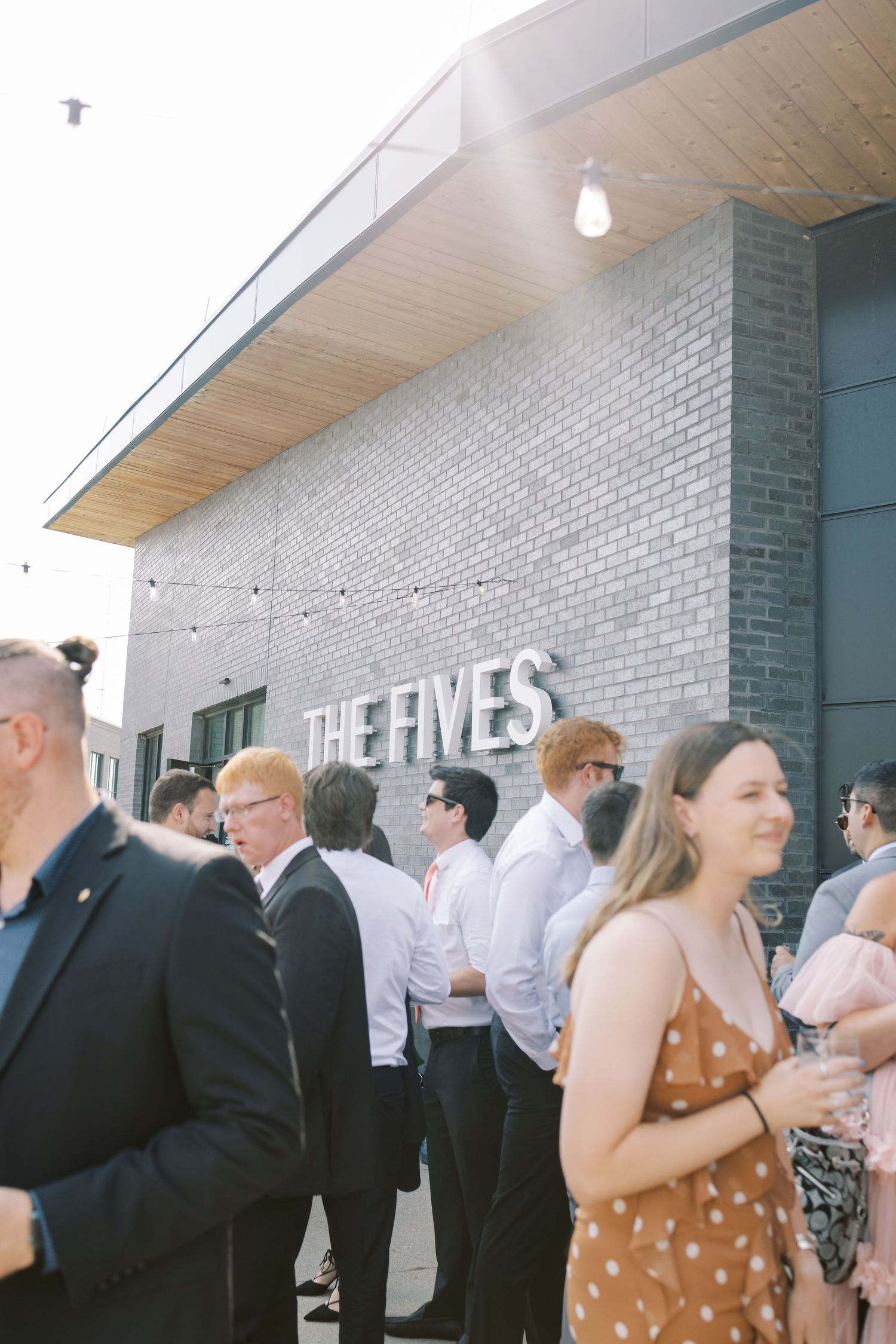 Wedding guests gather outside The Fives venue building with white brick walls and string lights overhead.