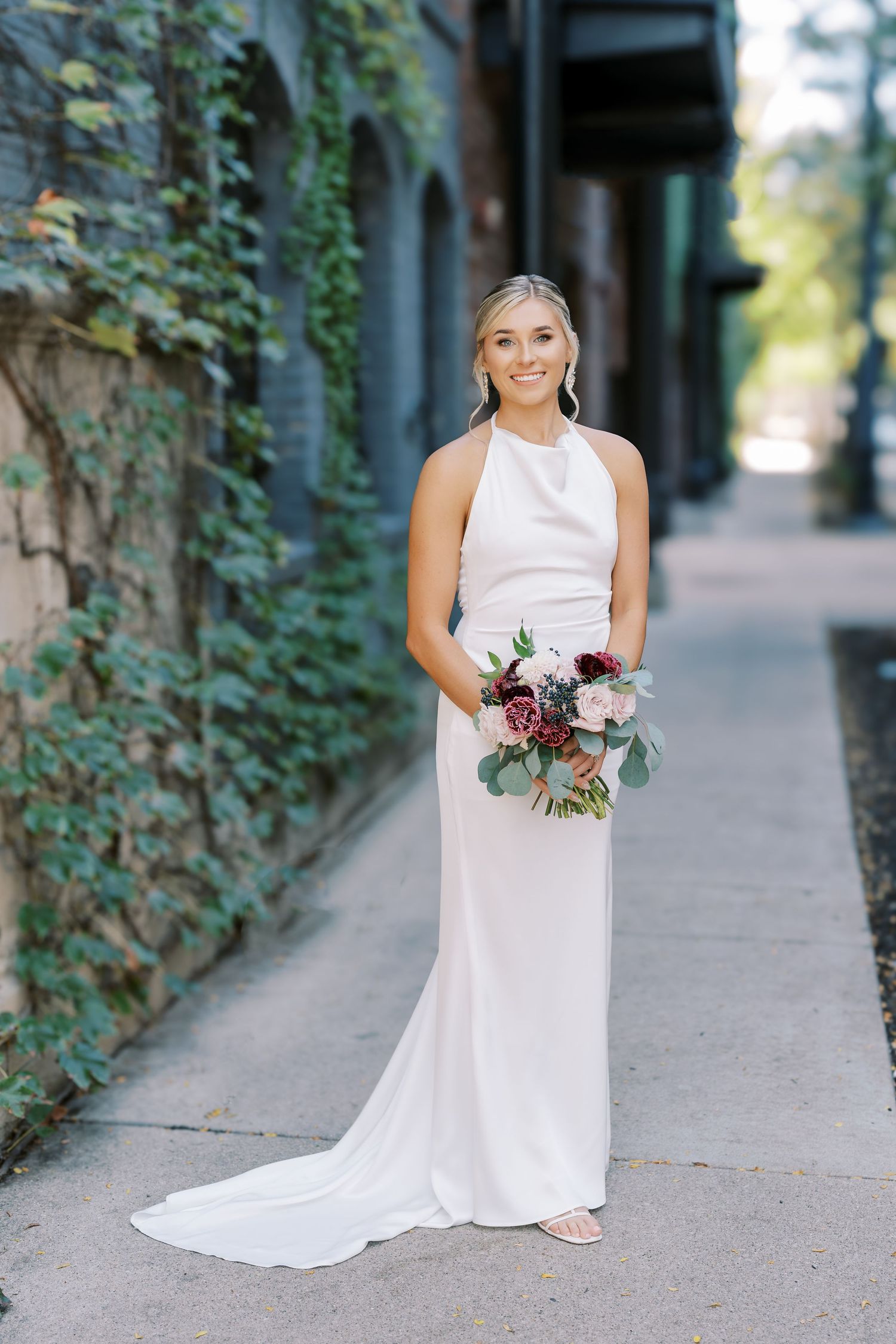 A bride in a sleeveless white gown poses along an ivy-covered wall in an outdoor setting.