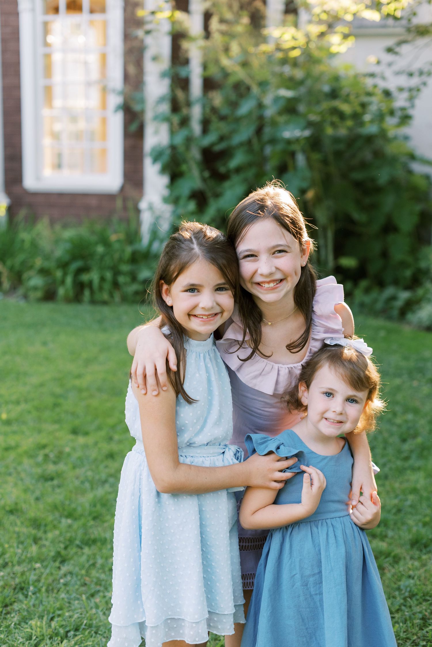 Three children embracing and smiling together on a grassy lawn.