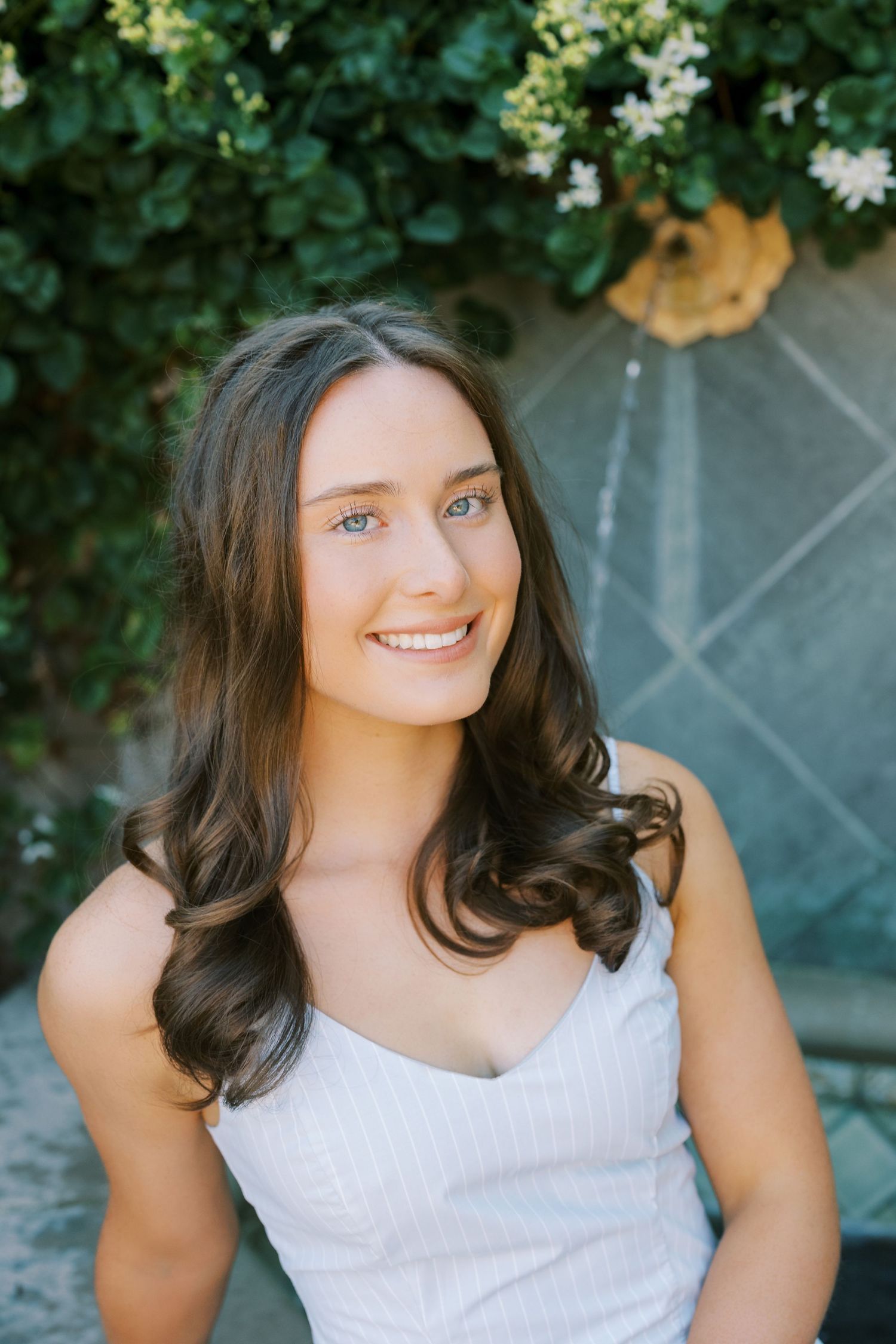 Portrait of someone in a white sleeveless top smiling outdoors with greenery and flowers in the background.