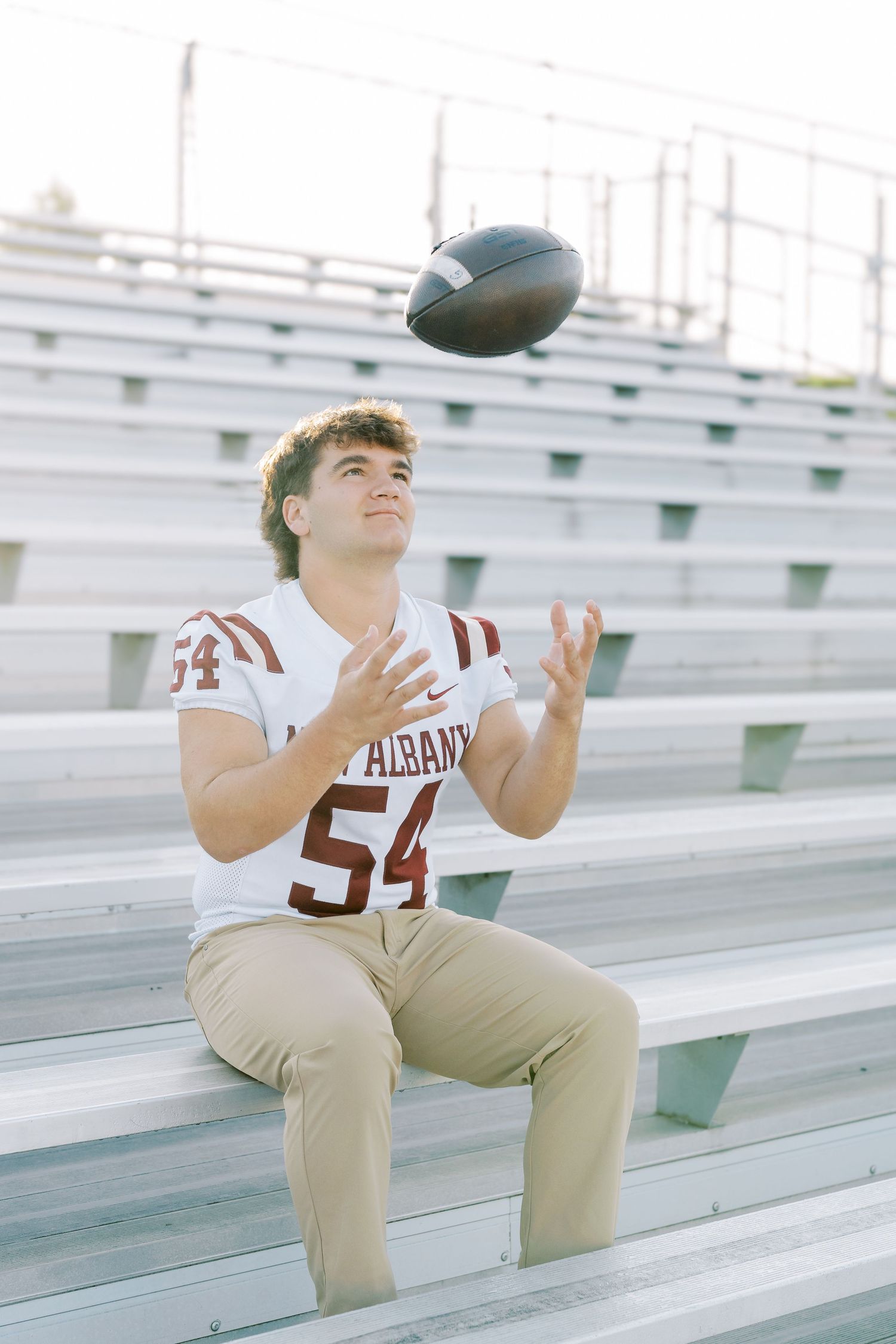 Football player in a jersey number 54 sits on stadium bleachers catching a football in multiple action poses.