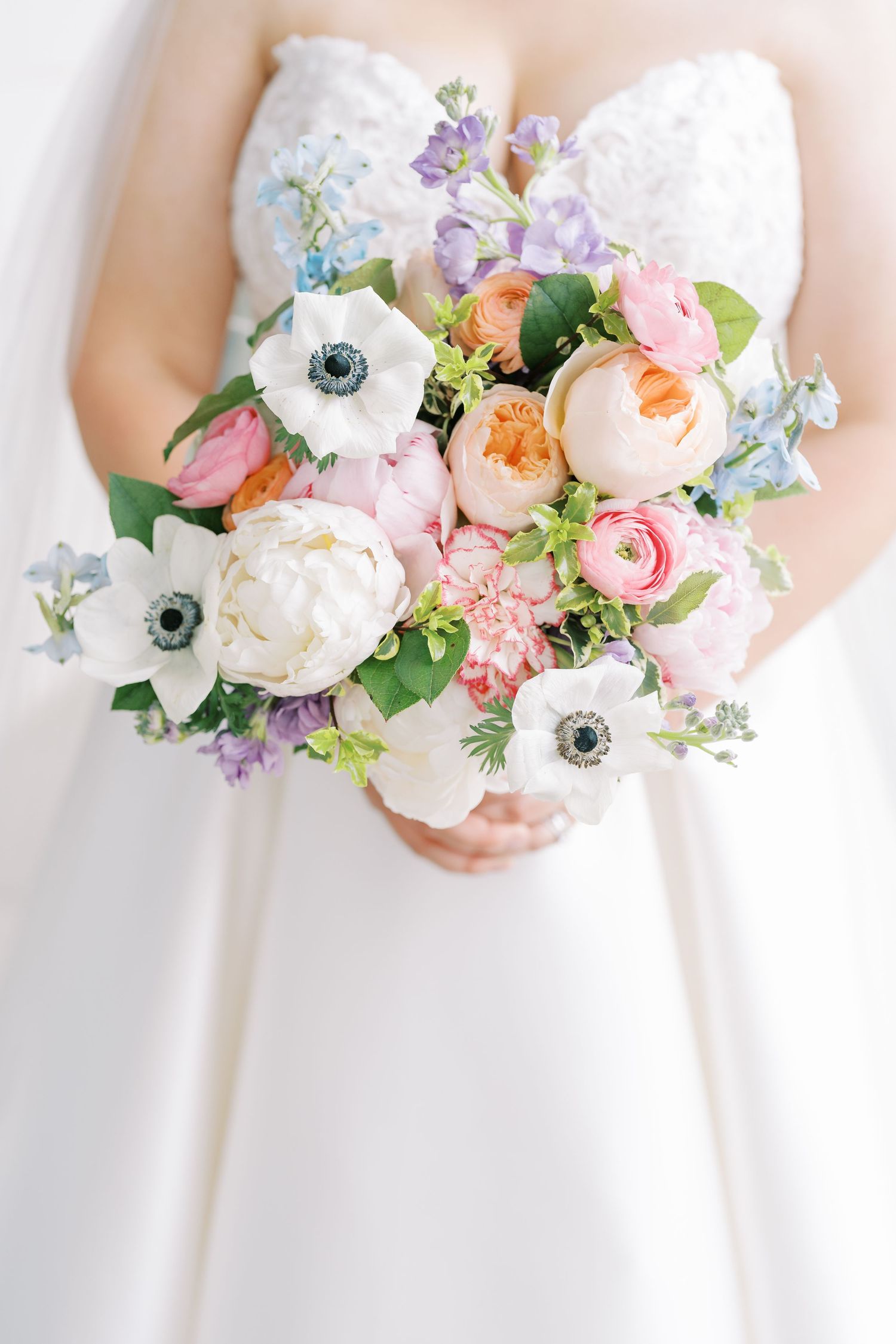 Bridesmaids in white dresses hold pastel bouquets with pink peonies, white anemones, and blue delphinium flowers.
