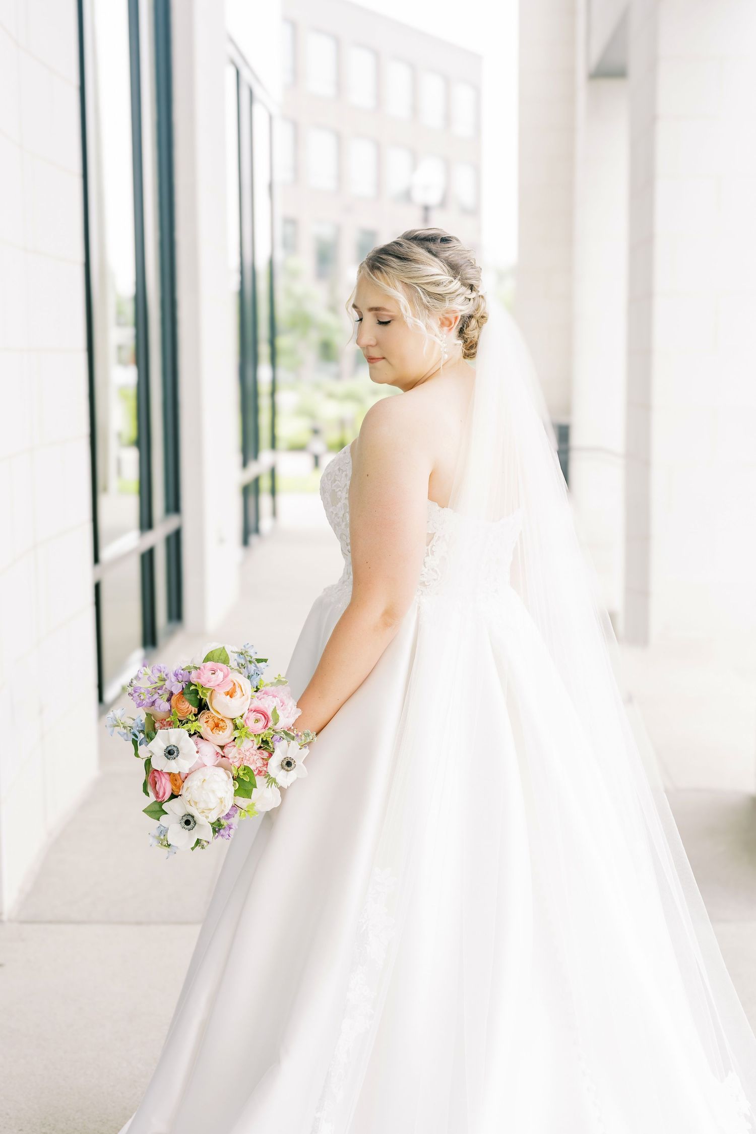 A bride in a flowing white gown holds a colorful bouquet of pink and white flowers in a bright sunlit hallway.