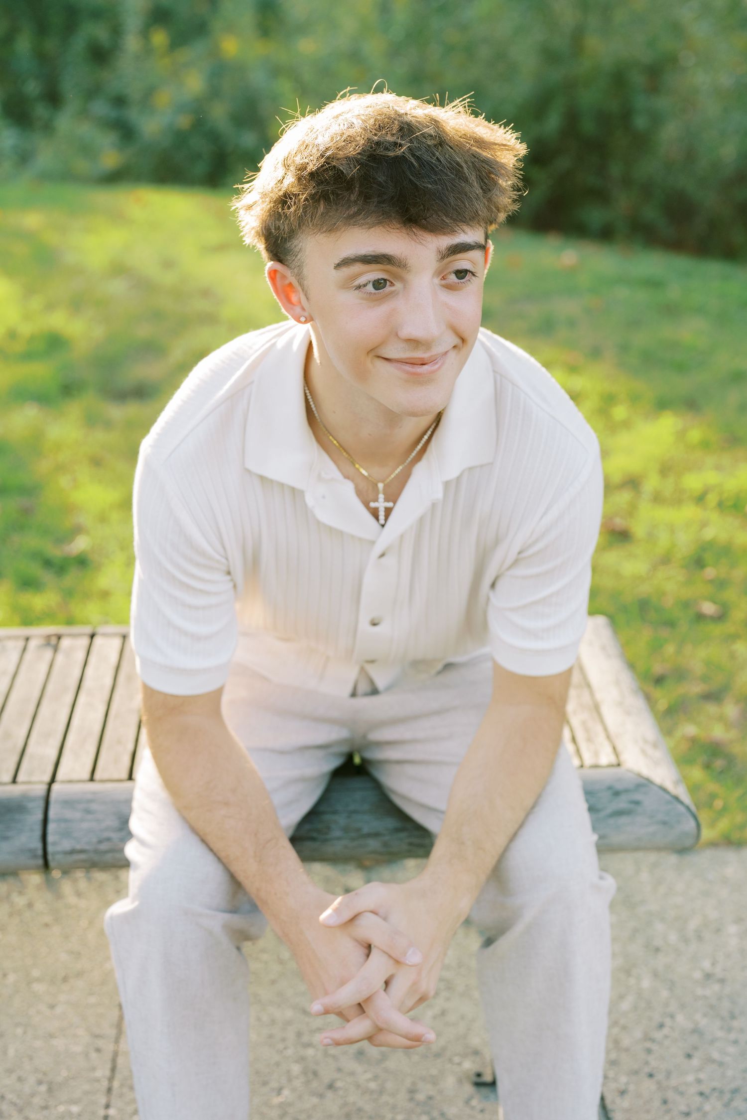 A person in white clothing sits casually on a wooden bench outdoors in a sunny garden setting.