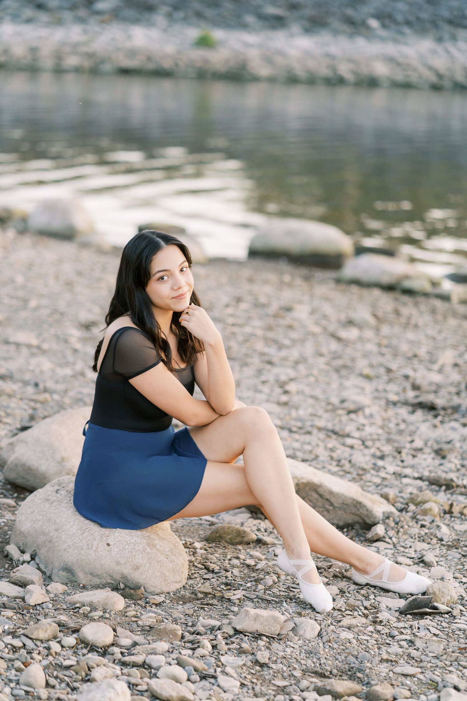 A person sits thoughtfully on rocky riverbank wearing casual summer attire and white socks.