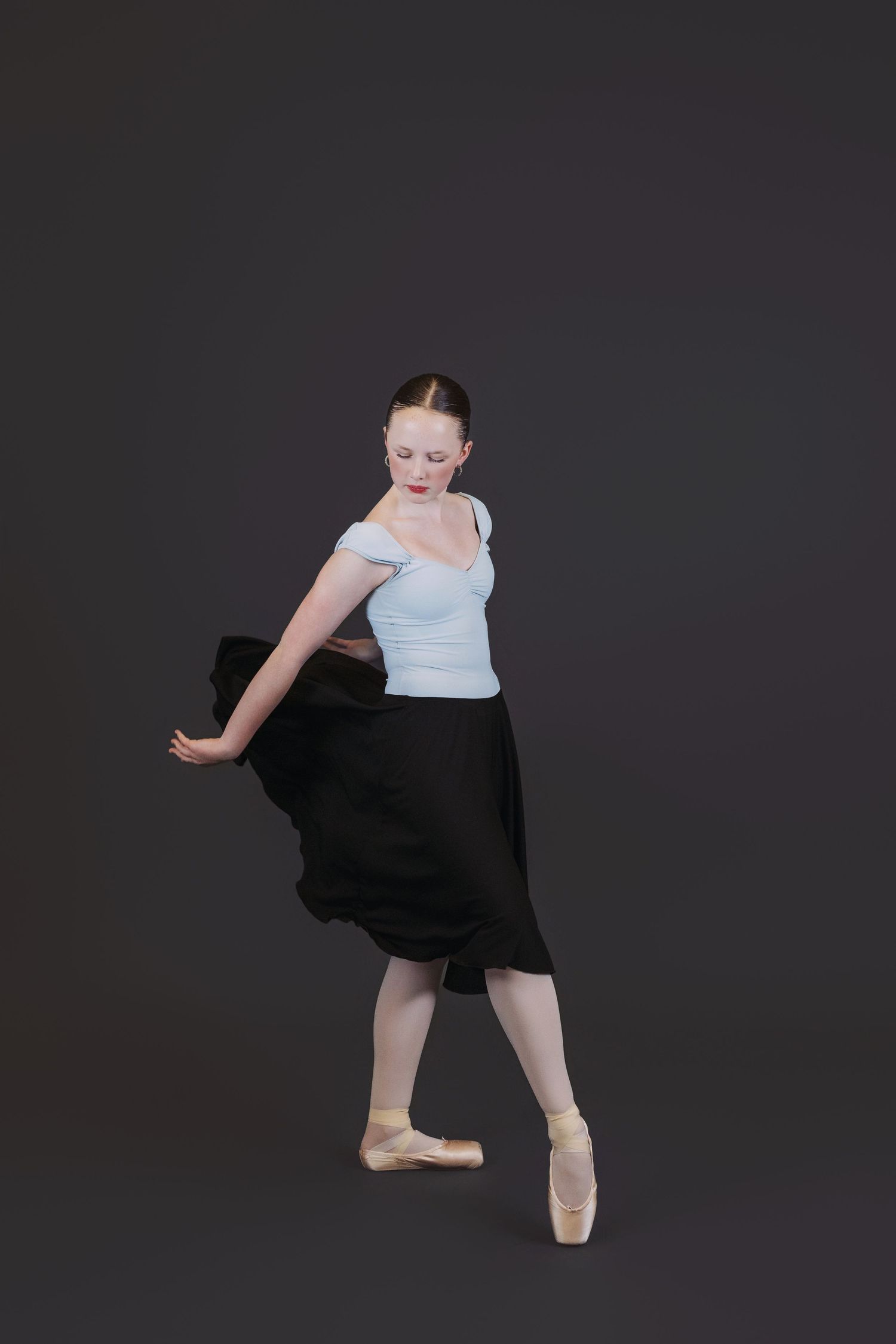 Ballet dancer in white top and black skirt performs graceful pose en pointe against dark background.