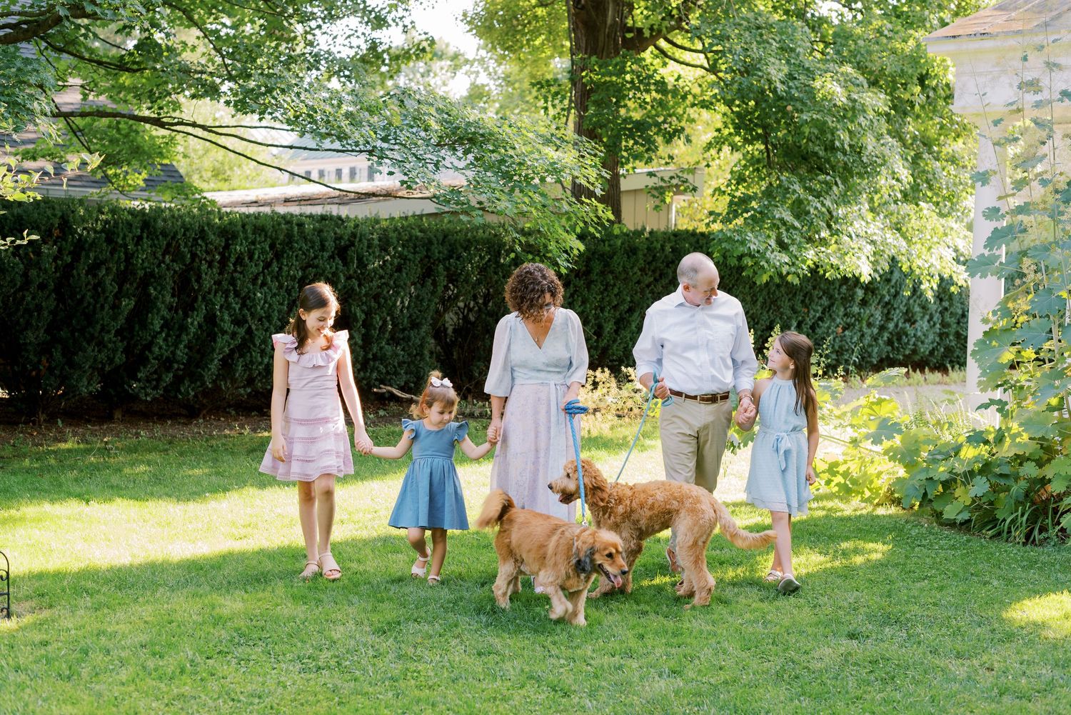 Family walking dog together on green lawn with hedges and trees in background.